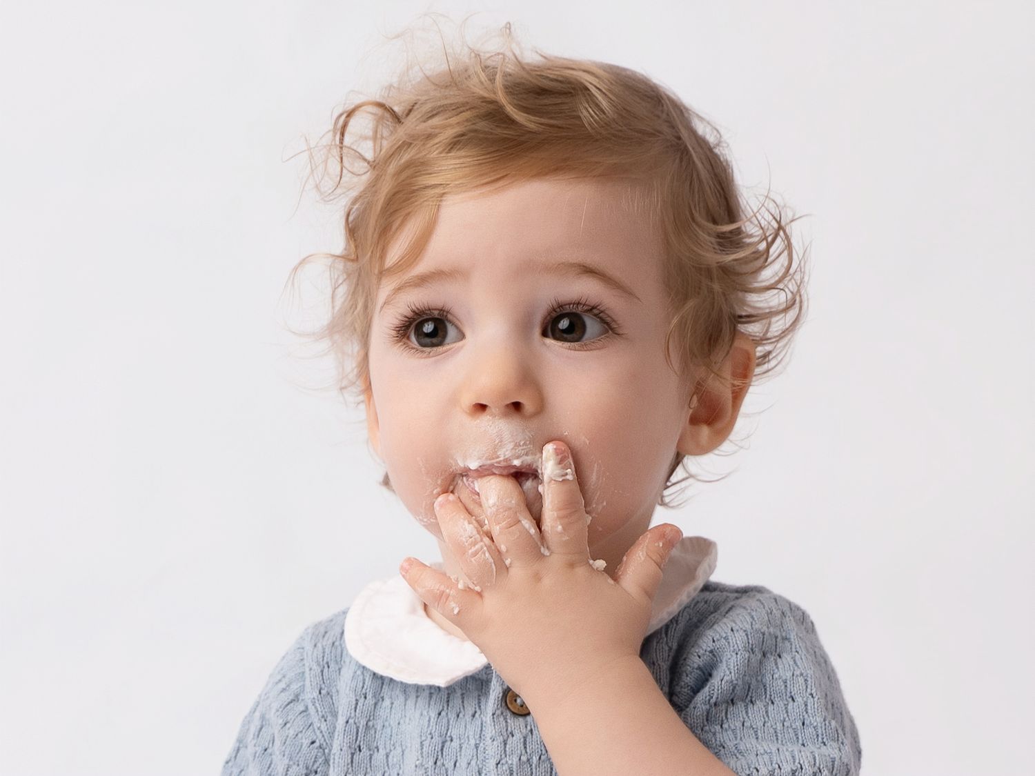Adorable toddler with curly hair covering mouth with frosting-covered hand against white background.