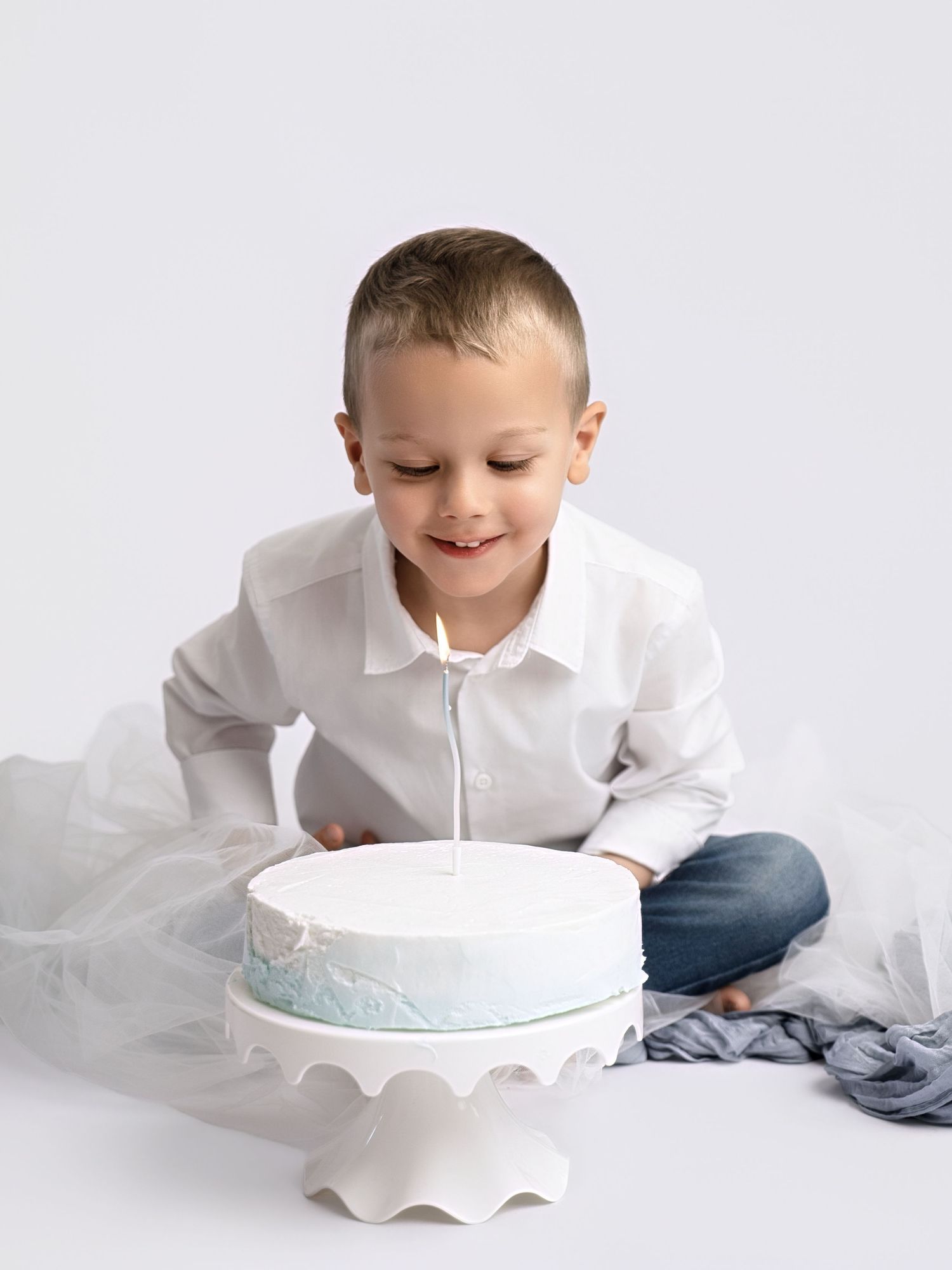 Young boy in white shirt smiling at birthday cake with candle on white cake stand.