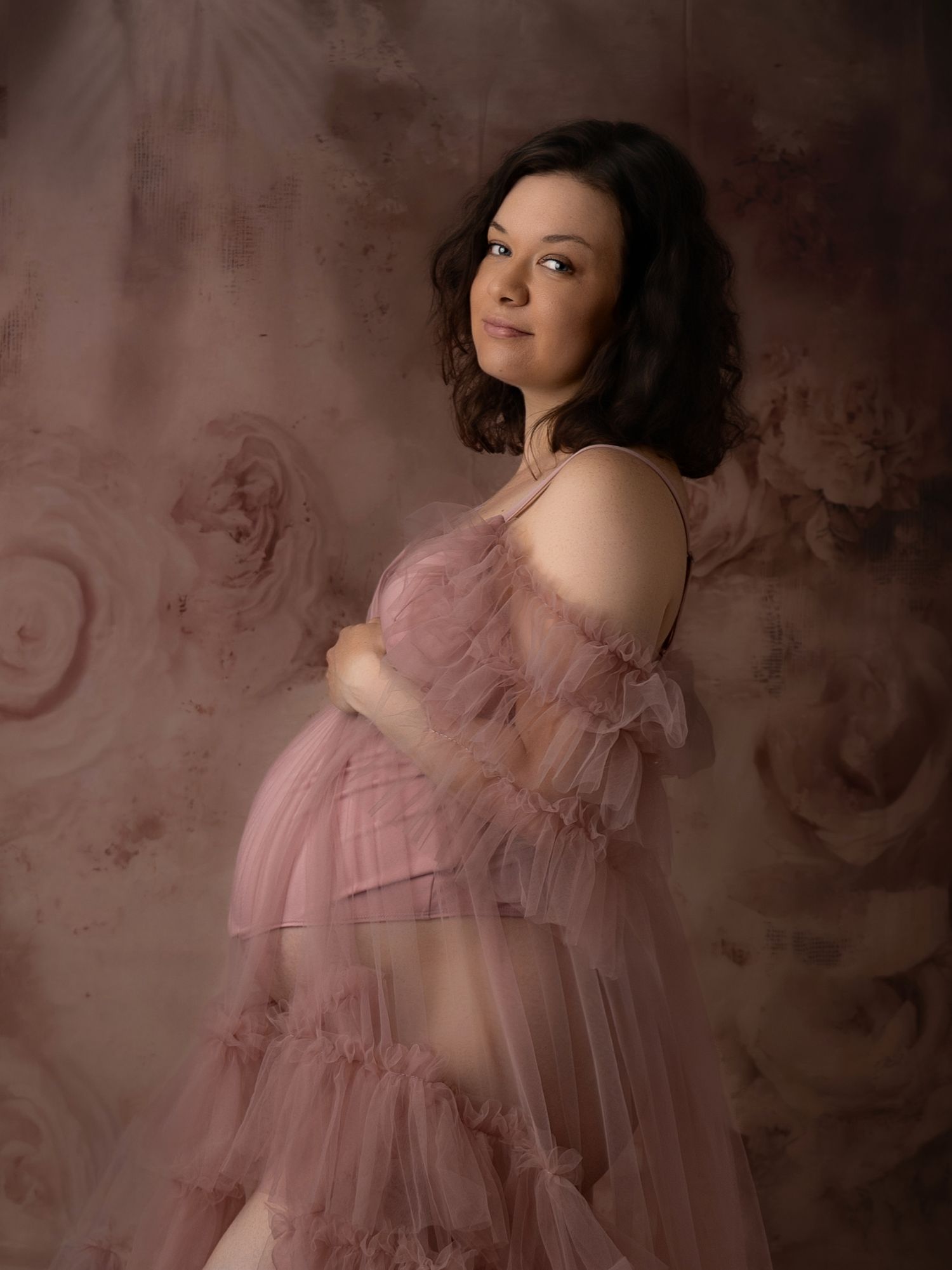 Pregnant woman in sheer pink tulle gown posing against floral backdrop in maternity shoot.