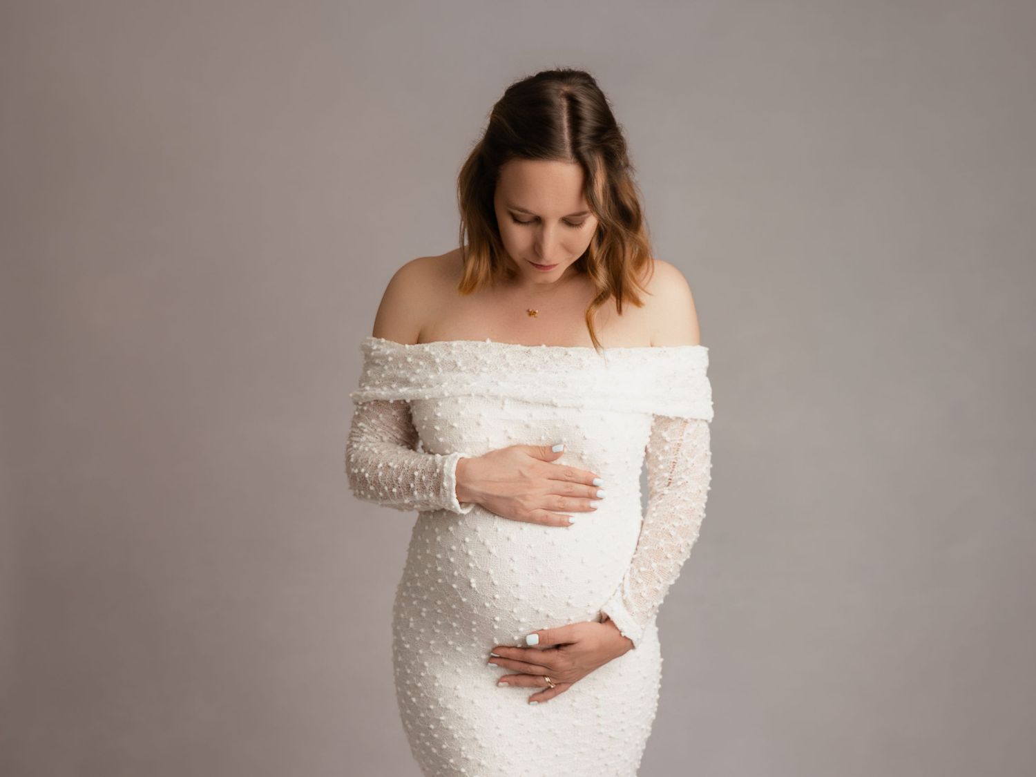 A pregnant woman in a white off-shoulder lace gown tenderly cradles her baby bump in a soft studio portrait.