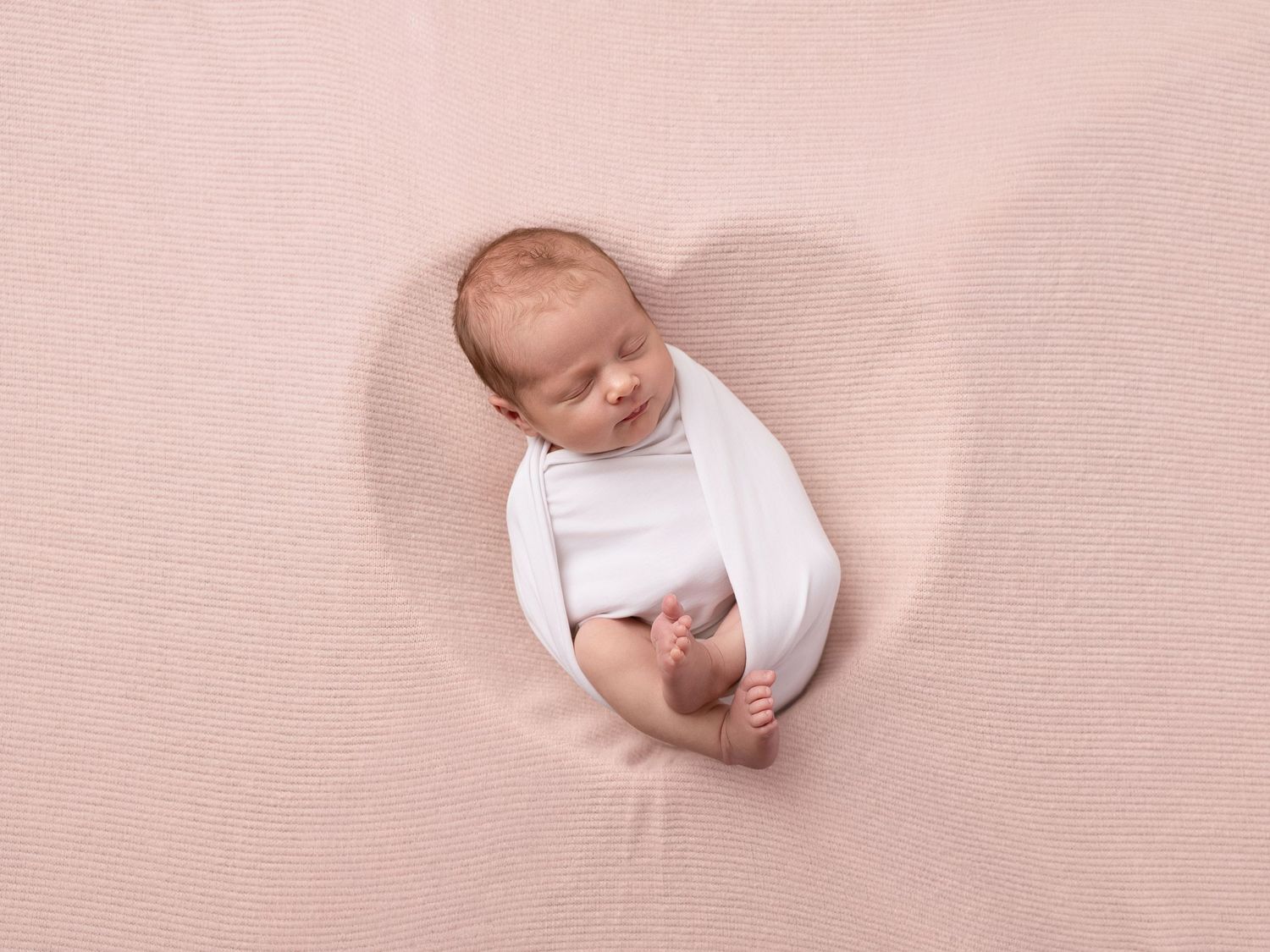 Newborn baby swaddled in white wrap sleeping peacefully on a pink heart-shaped blanket backdrop.