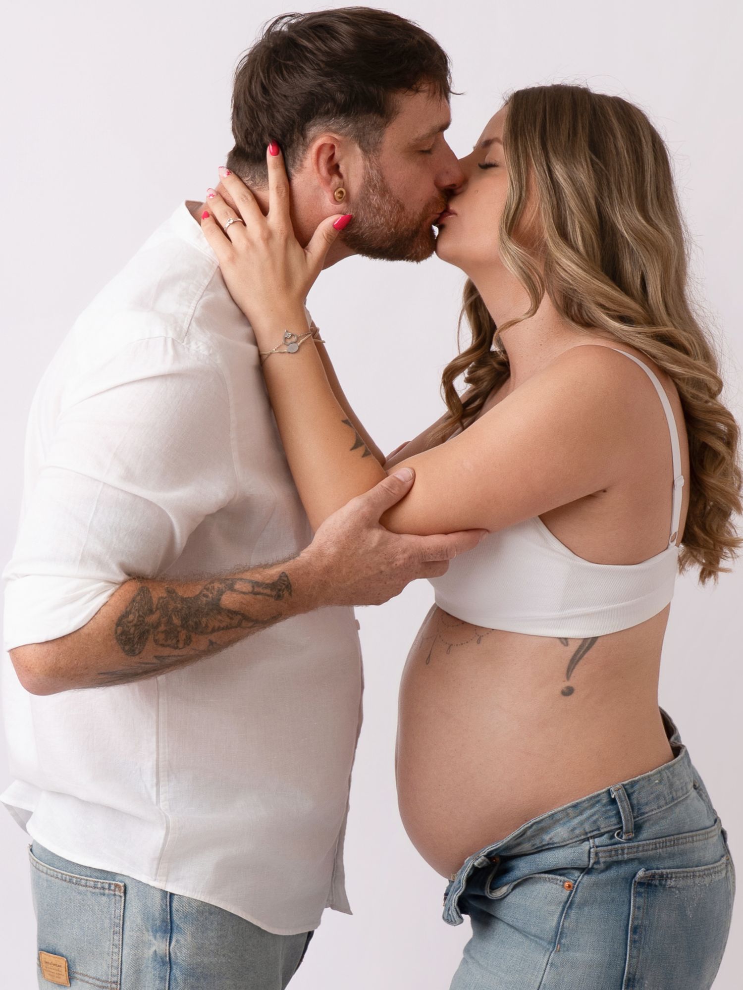 Couple sharing a kiss during maternity photoshoot, pregnant woman in white crop top and jeans, man in white shirt.