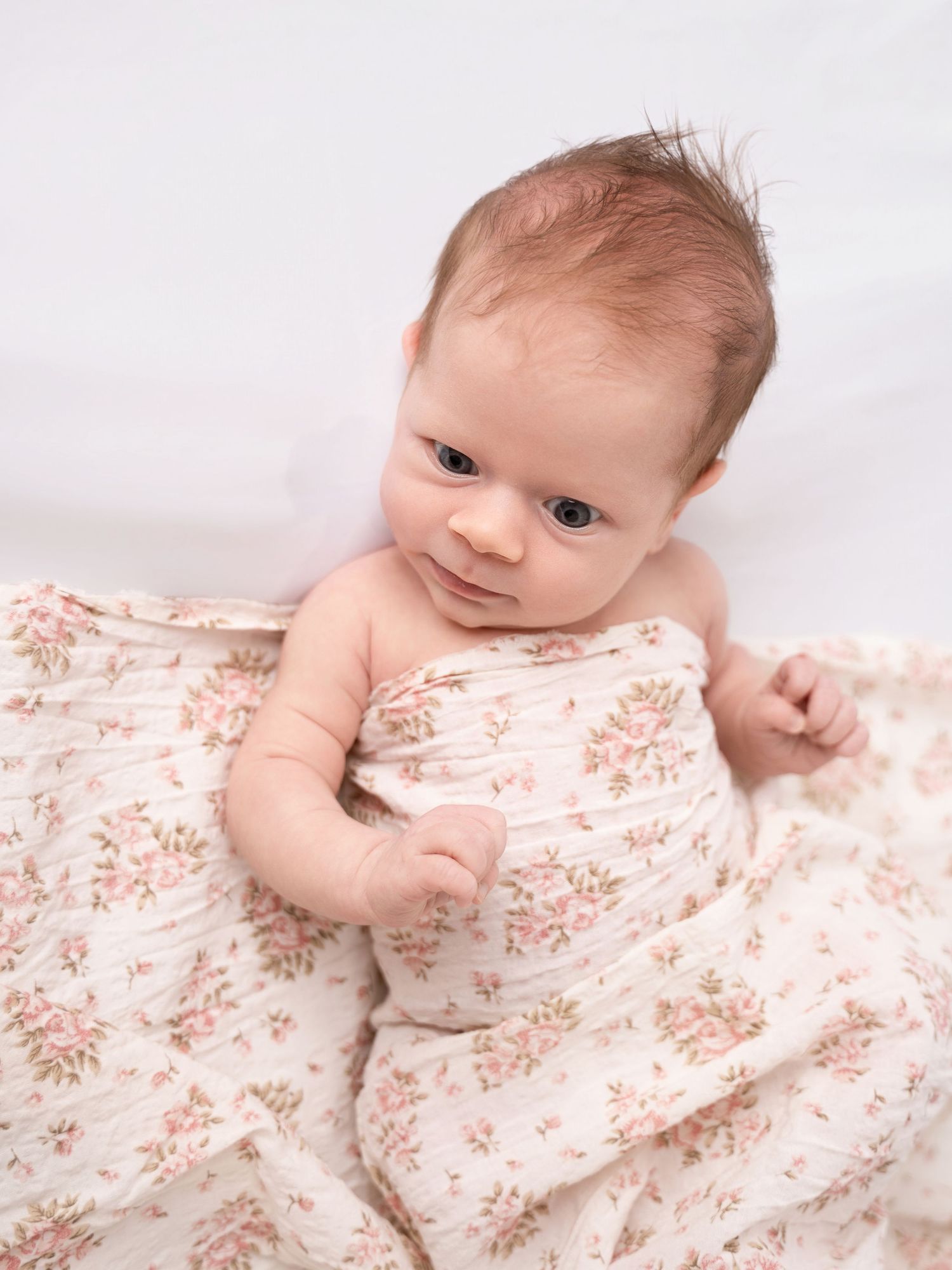 Newborn baby wrapped in a floral muslin swaddle blanket, looking up with bright eyes on a white background.