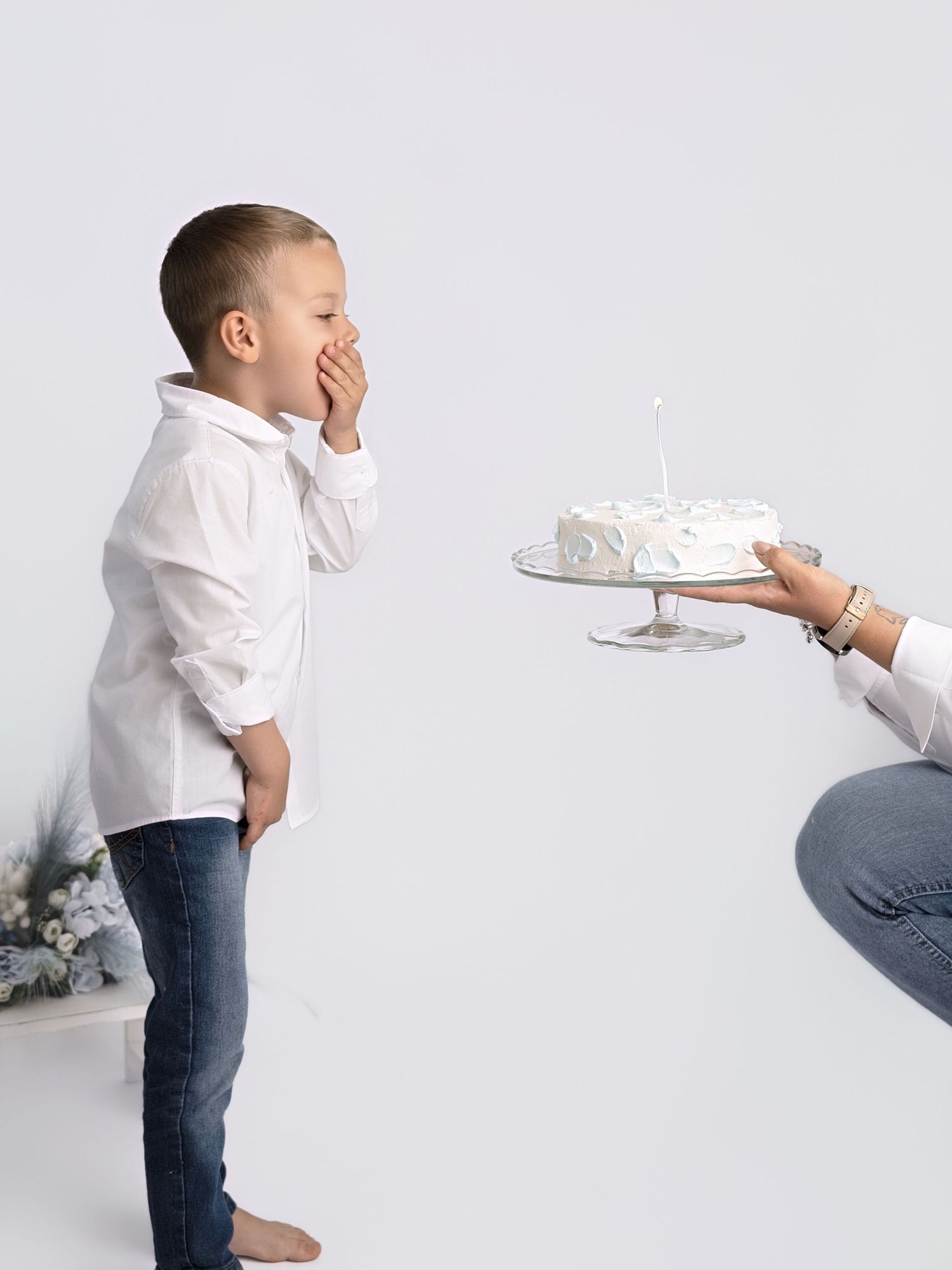 A young boy in a white shirt reacts with surprise as someone presents him with a white birthday cake with a candle.