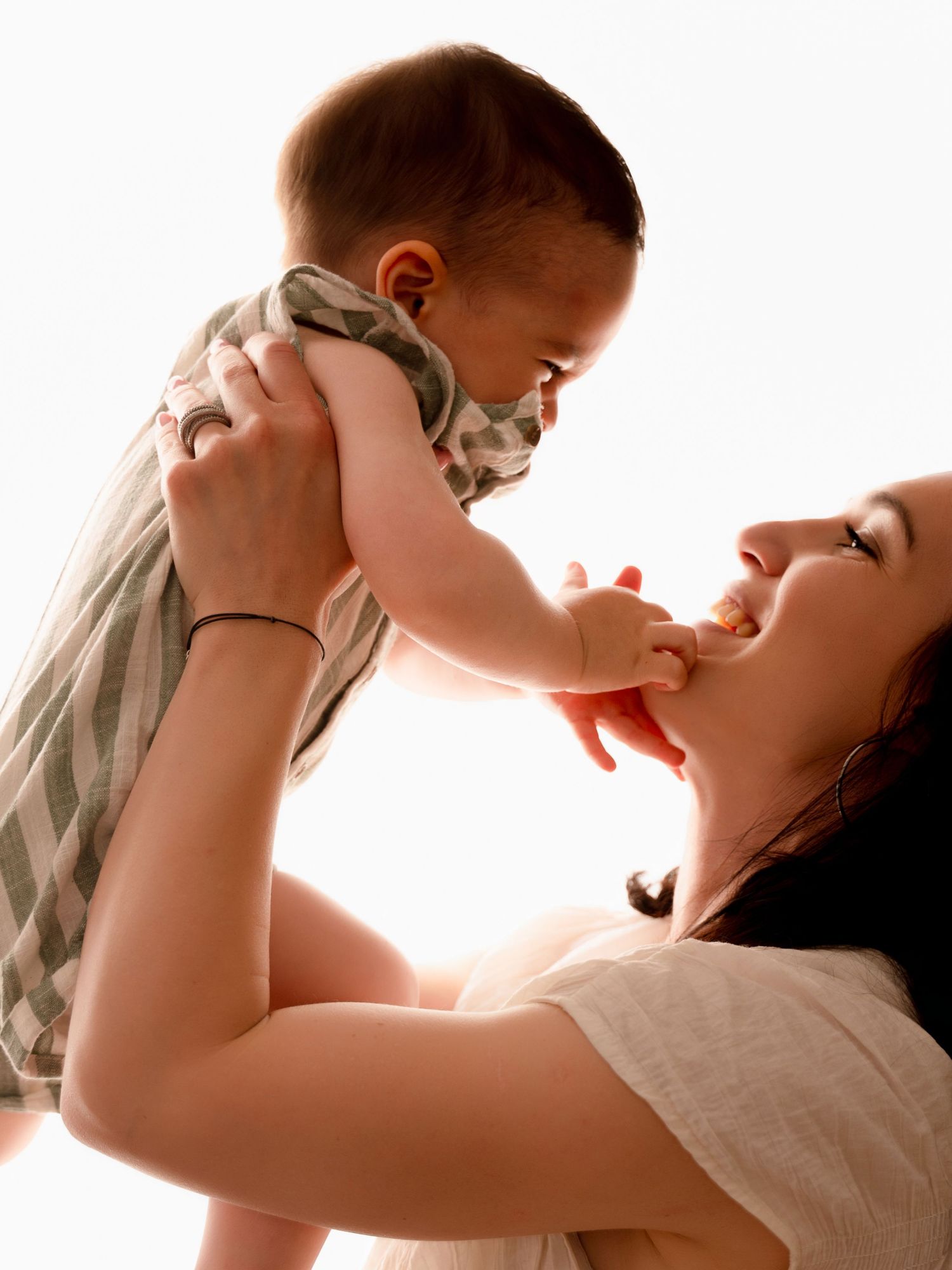 A joyful mother lifts her smiling baby up in the air, sharing a tender moment in soft backlit light.