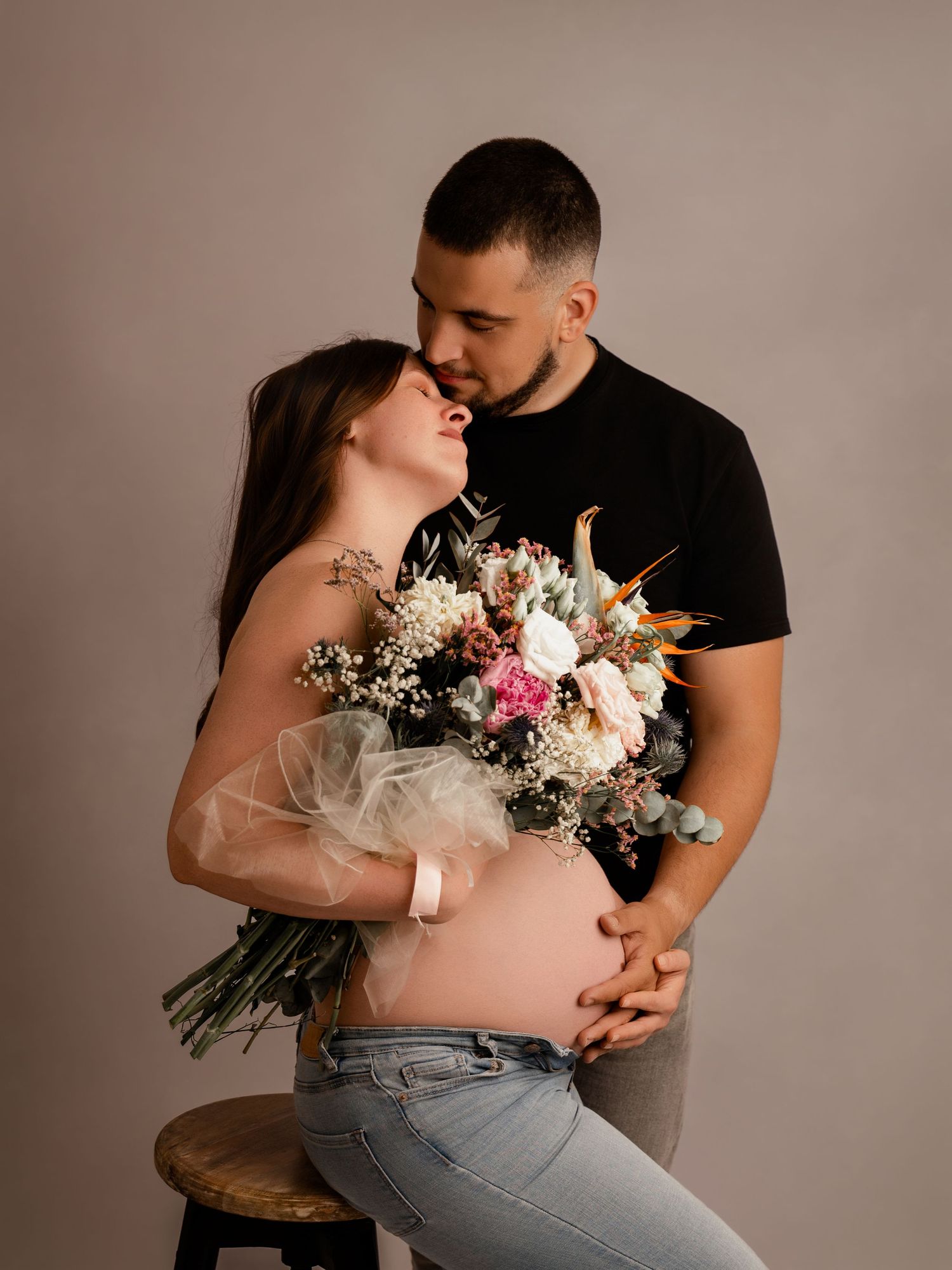Pregnant woman sitting on stool with floral bouquet as tattooed partner in black tee leans in to kiss her.