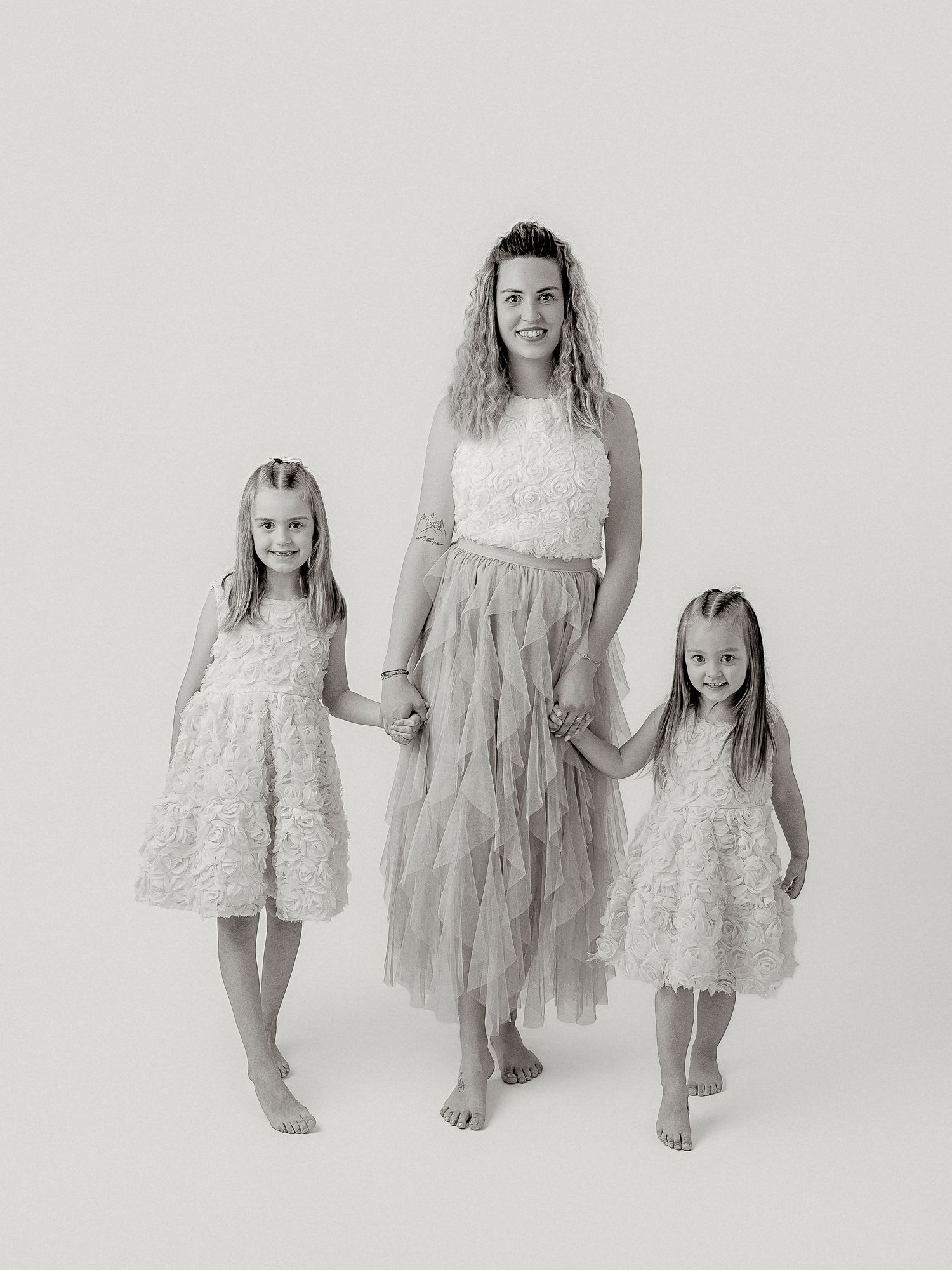 Black and white studio portrait of a mother holding hands with two young daughters in matching floral dresses.
