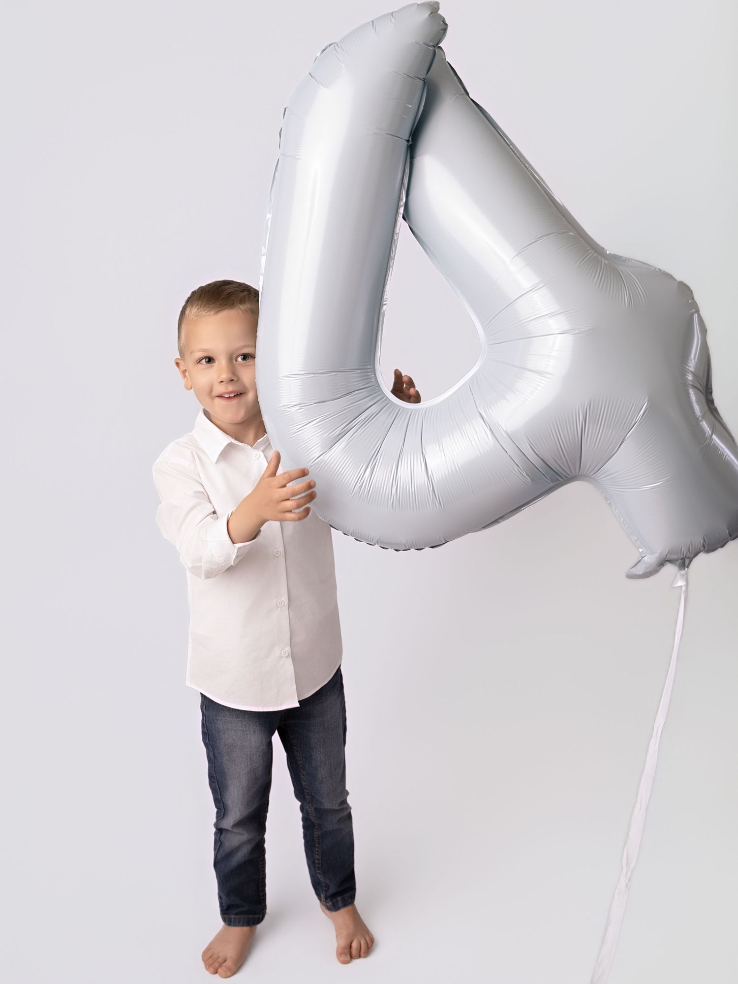 A young boy in a white shirt and jeans holds a large silver number 4 balloon, standing barefoot on a white background.