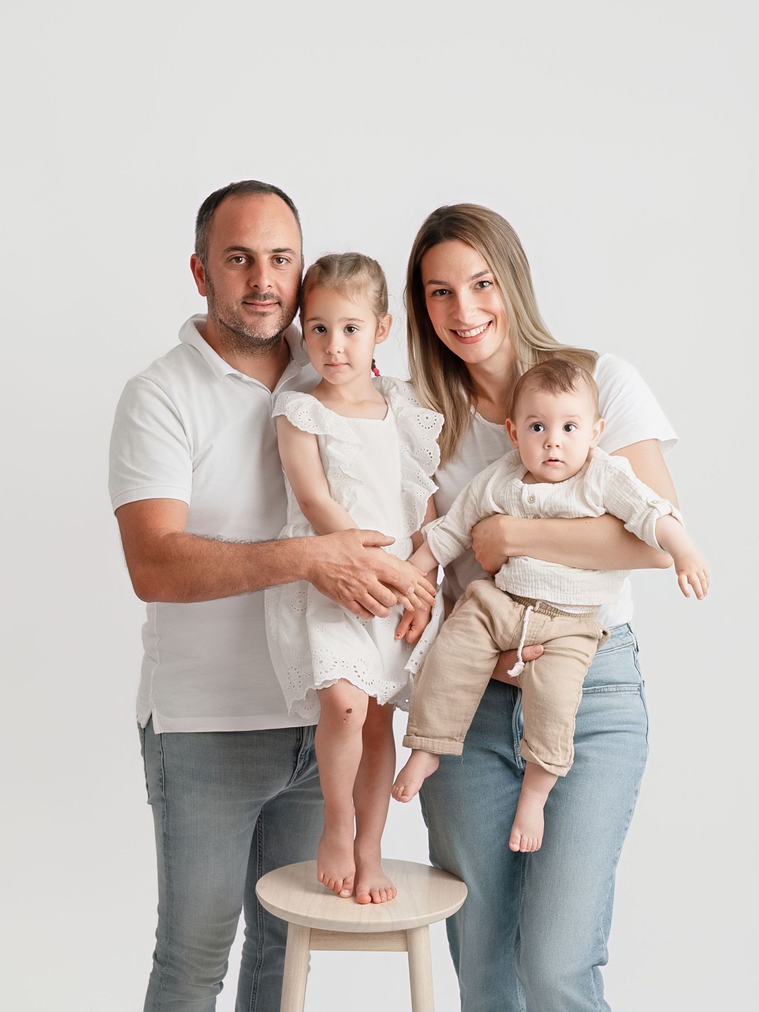 Family of four posing in white outfits against a white background, with two young children in a professional studio portrait.