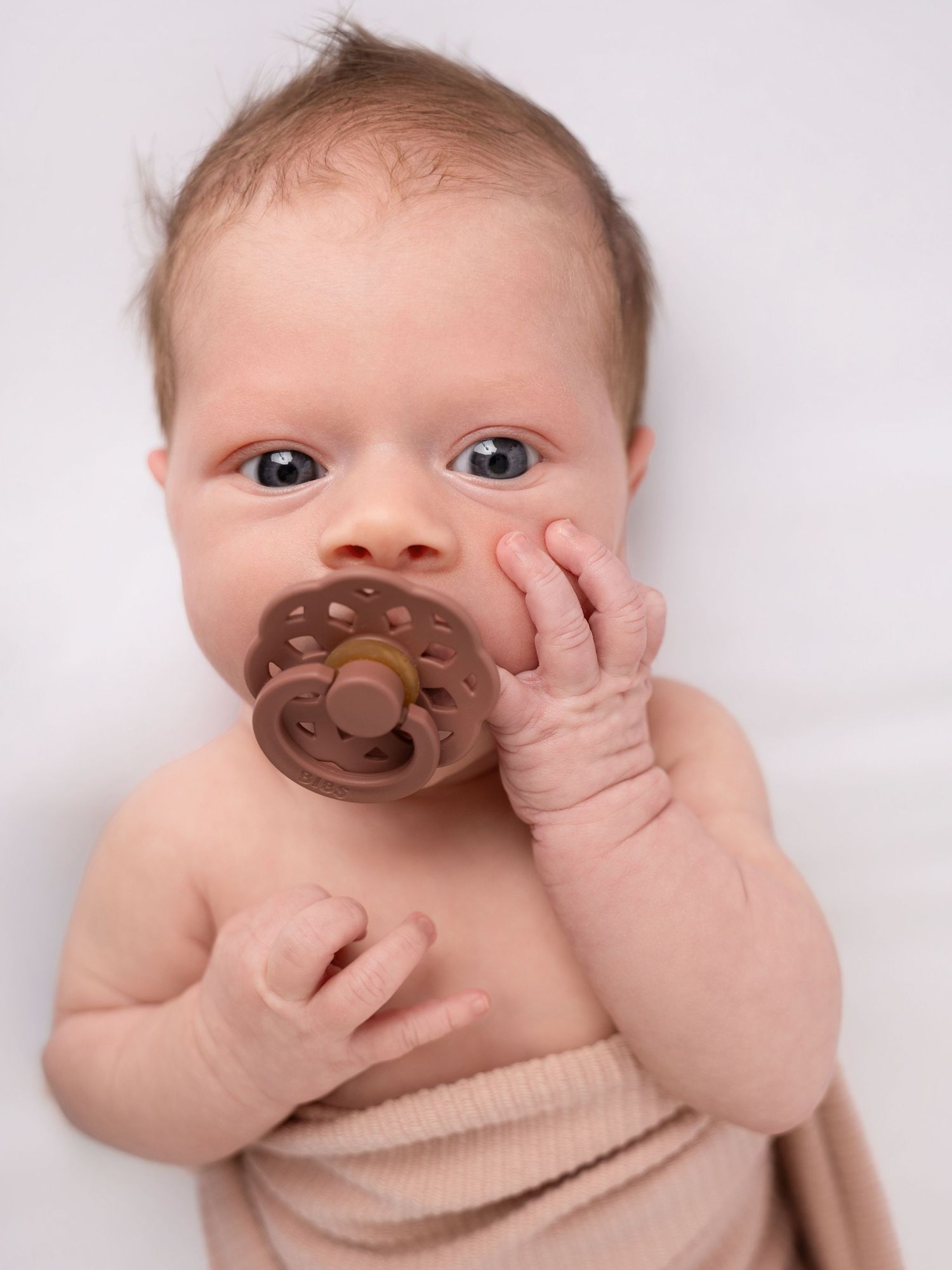 A curious baby with blue eyes holds a brown pacifier while lying on a white blanket.