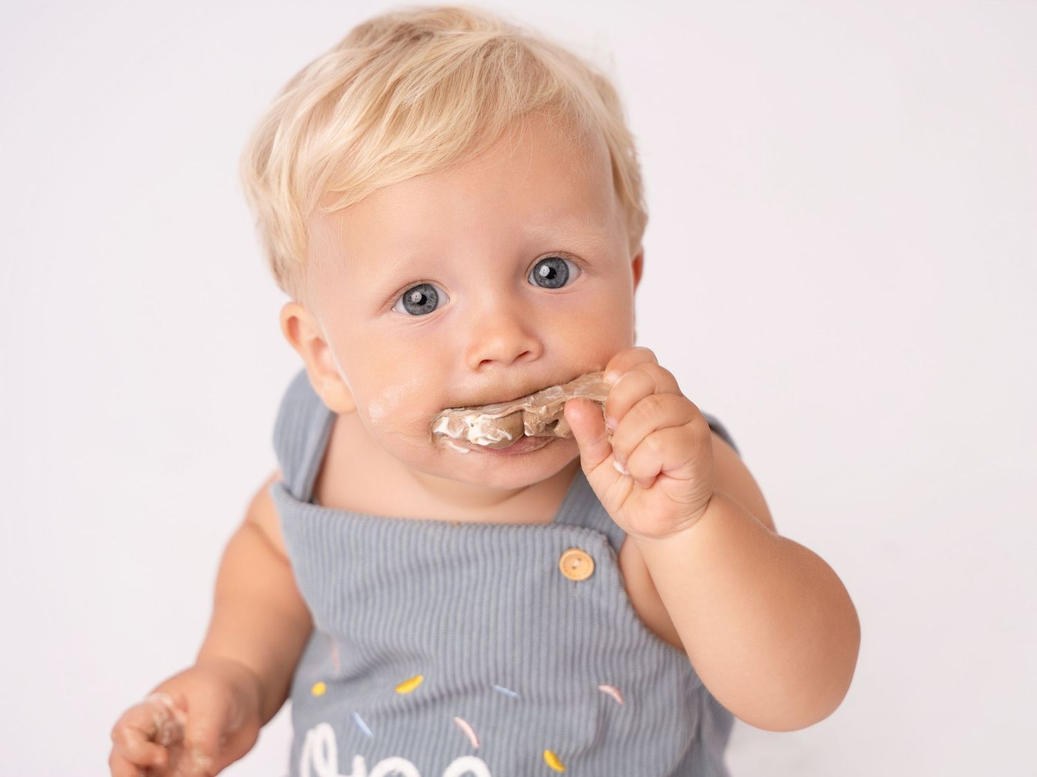 Cute blonde toddler in gray overalls eating a cookie, blue eyes, white background, baby photo.