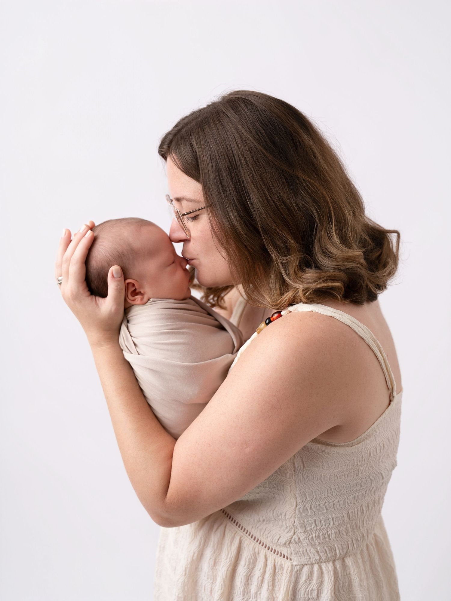 A mother in a cream dress gently touches noses with her swaddled newborn baby.