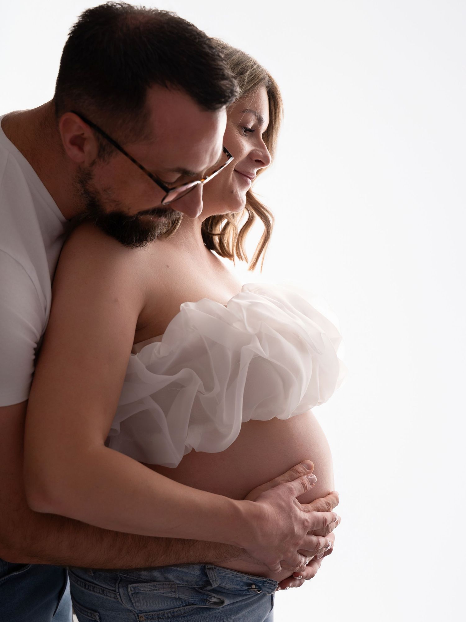 A couple shares a tender maternity photo, man embracing pregnant woman in white tulle top and jeans against white background.