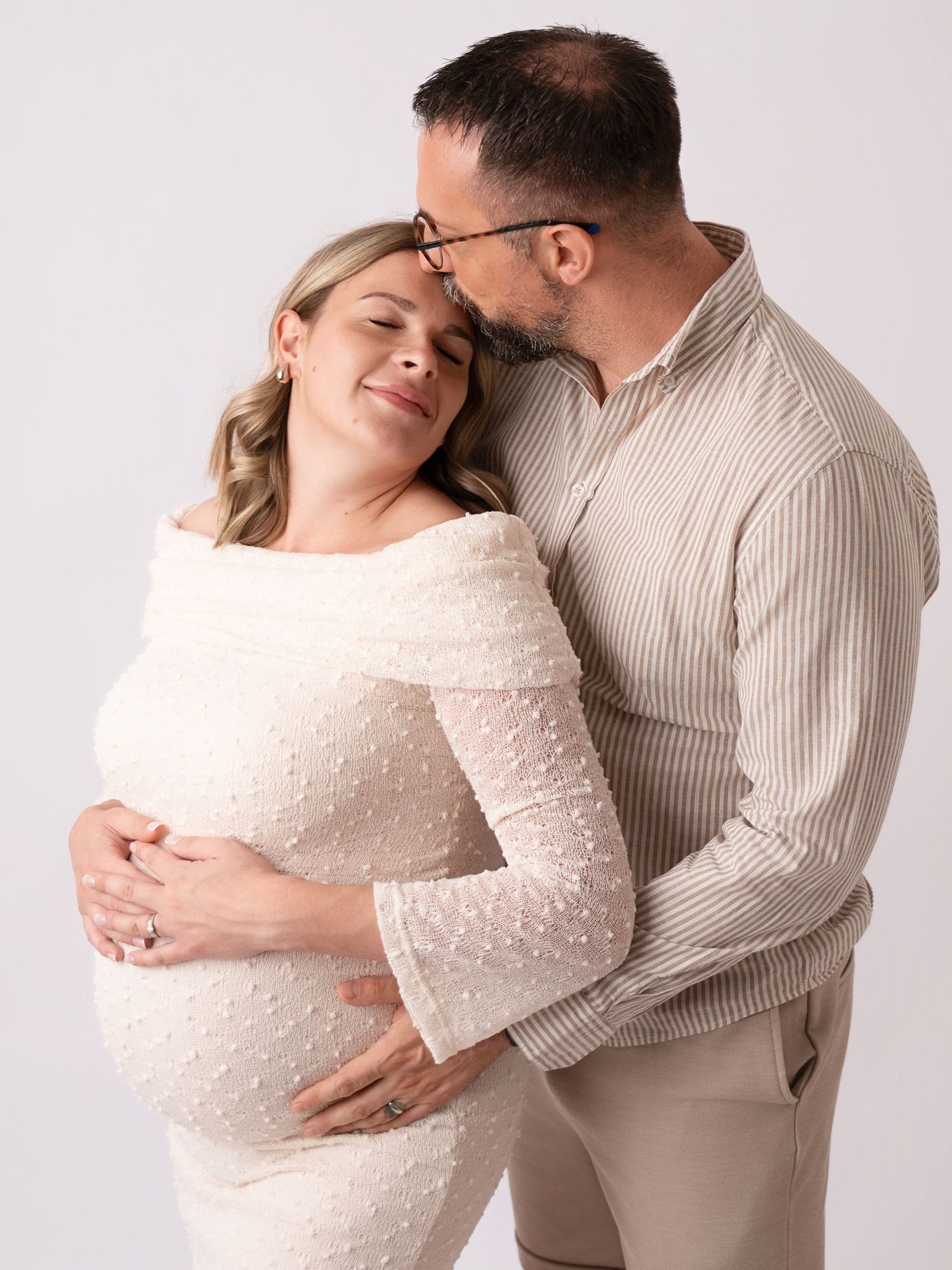Couple sharing a tender maternity photo shoot moment, man kissing pregnant woman's temple while cradling her belly.