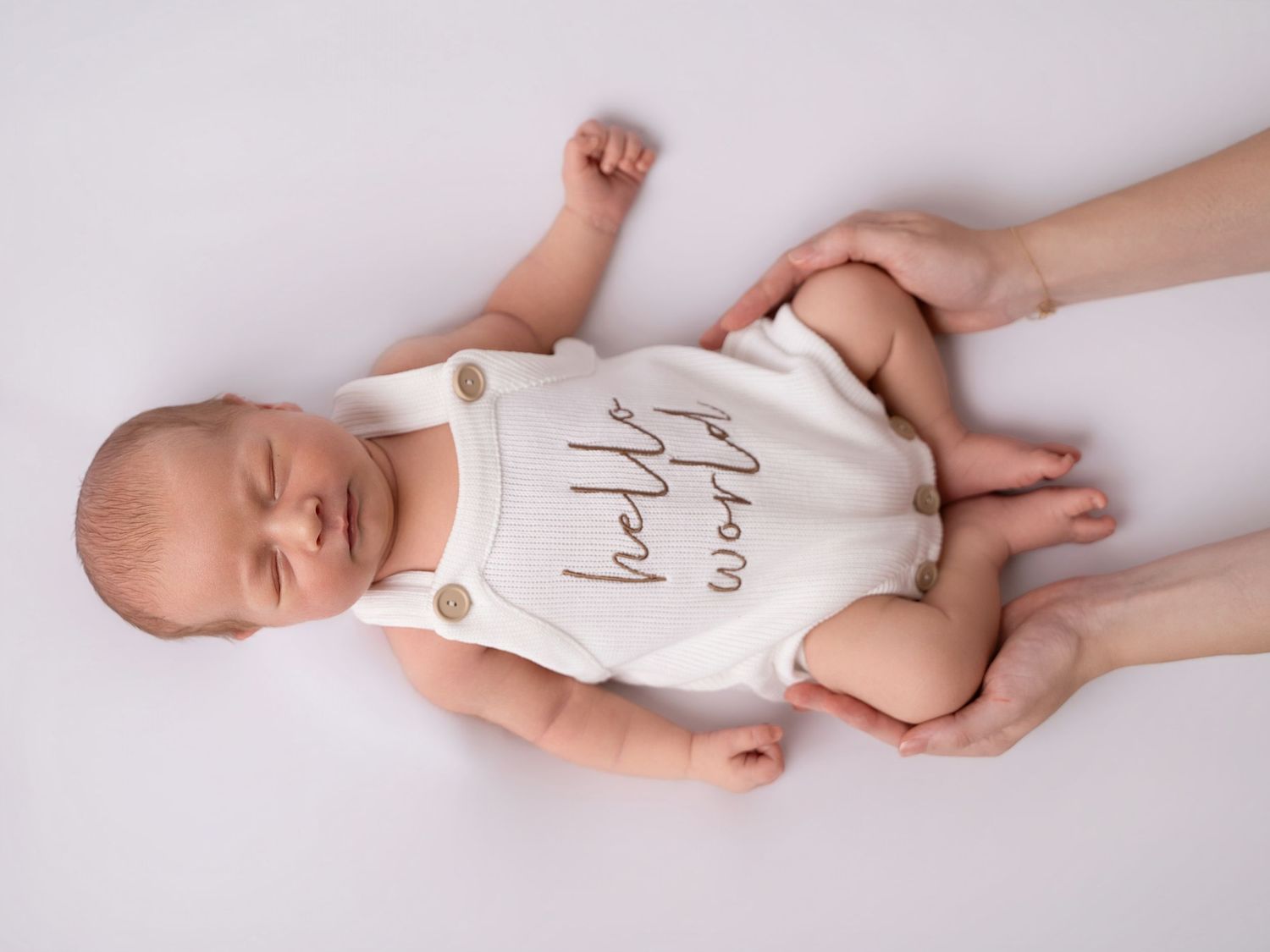 Newborn baby in white Hello World onesie held by adult hands on white background.
