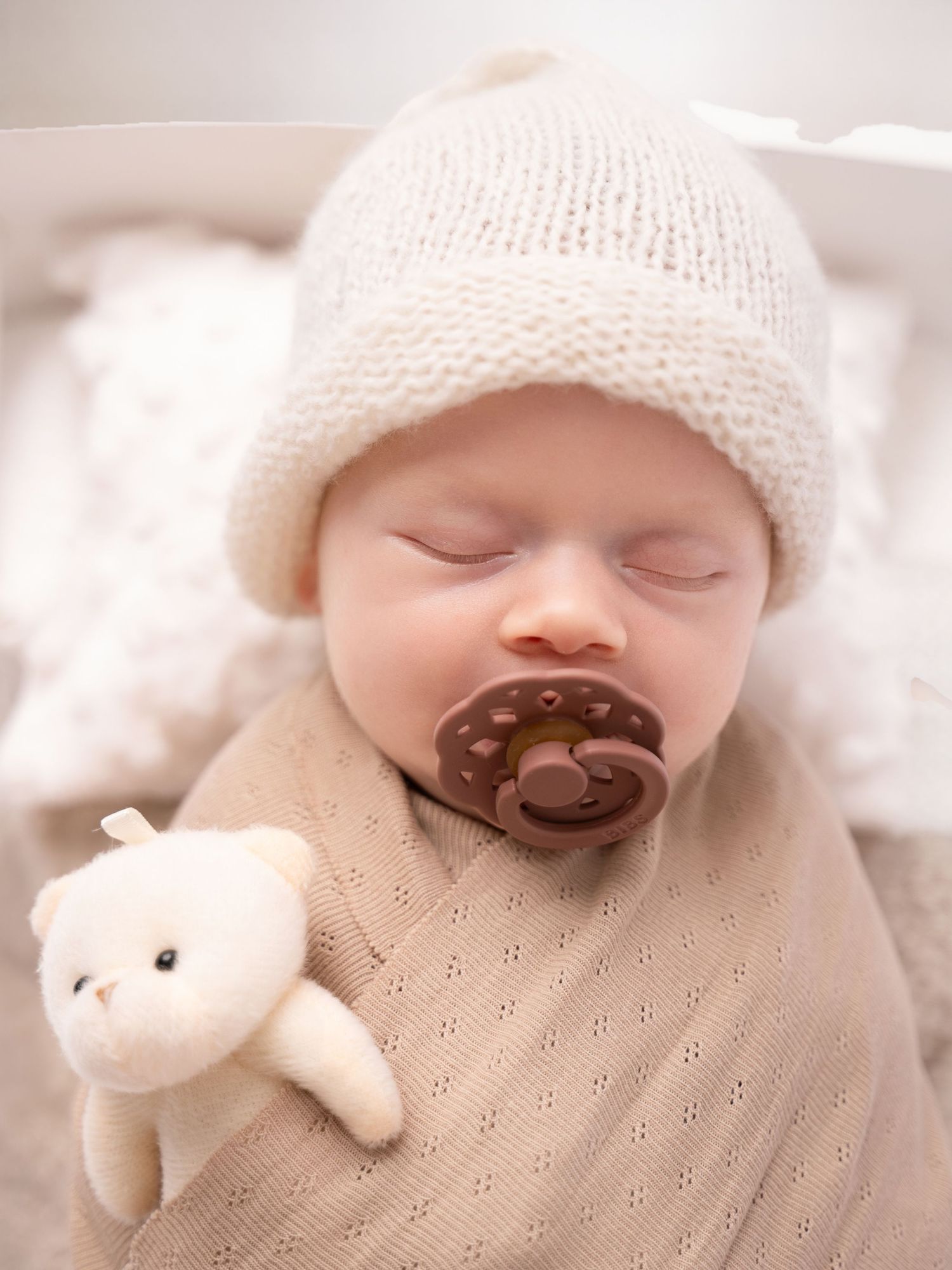 Sleeping newborn baby wearing a cream knit hat with a brown pacifier, swaddled in a beige blanket with a white teddy bear.