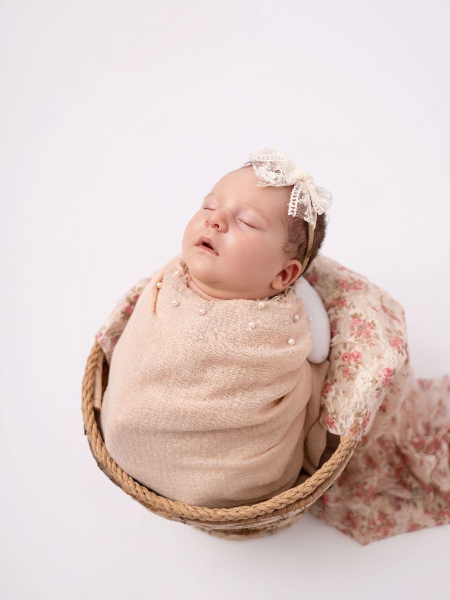 Sleeping newborn baby swaddled in a beige wrap with a lace bow headband, nestled in a wicker basket.