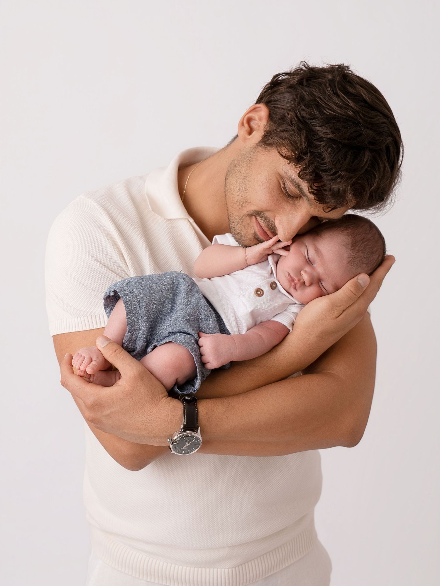Young father in cream shirt tenderly holds sleeping newborn baby, studio portrait on white background.