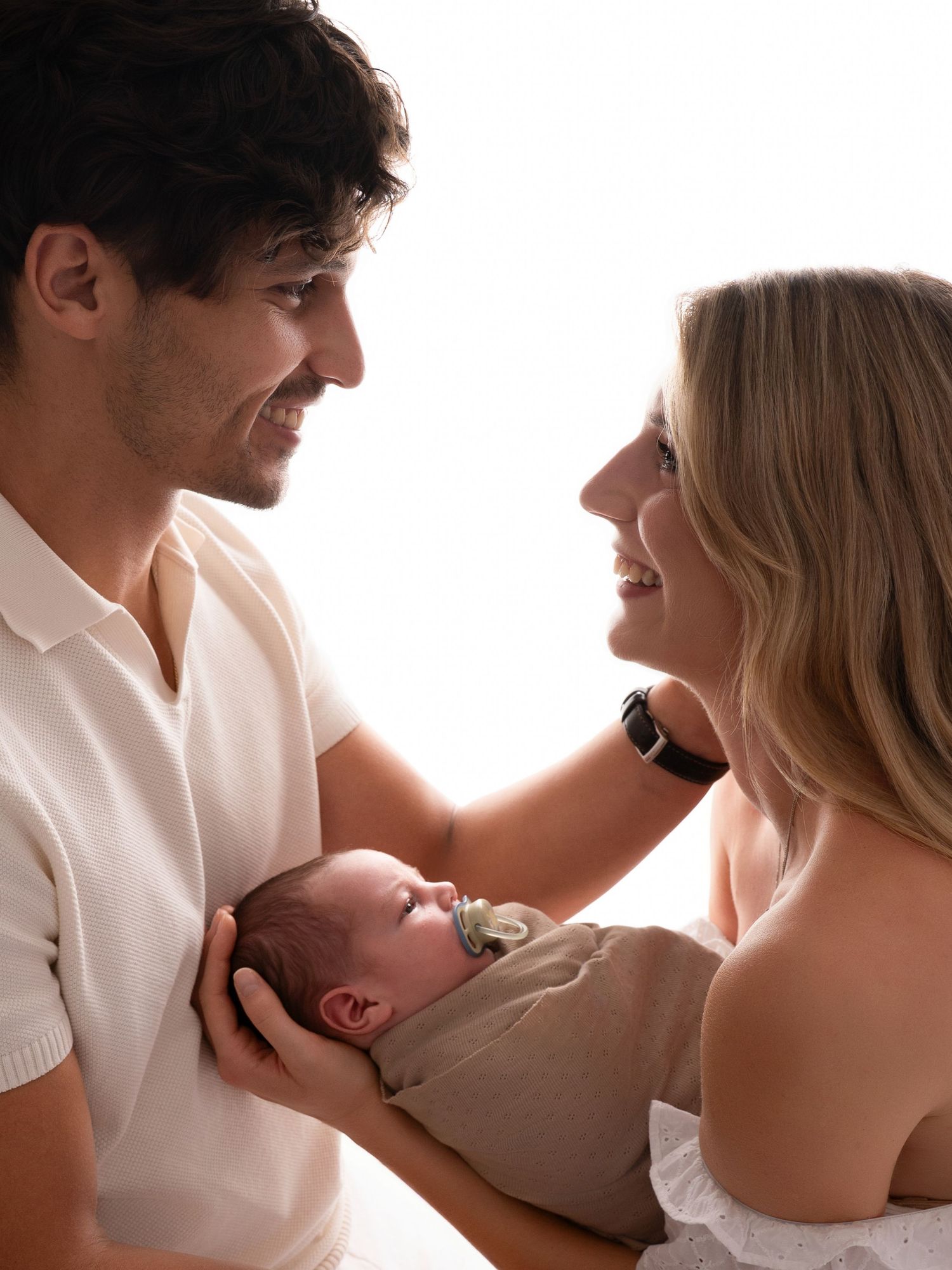 A smiling couple gazes lovingly at their newborn baby wrapped in a beige swaddle in a bright studio setting.