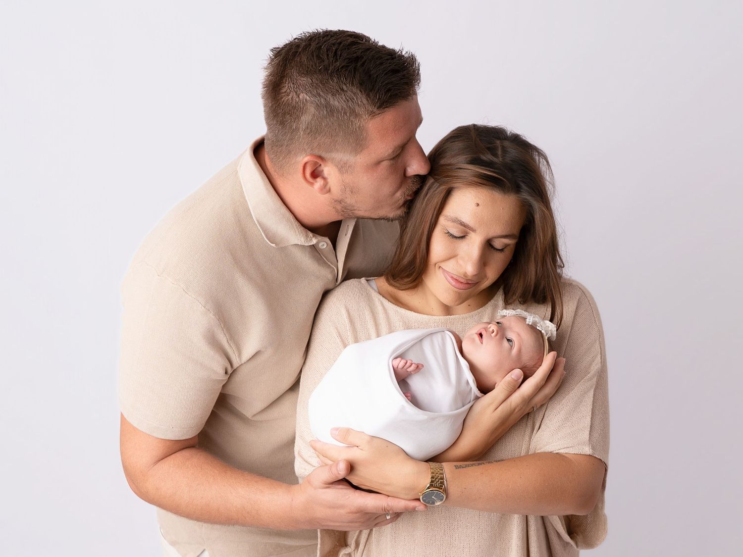 A couple in matching beige outfits tenderly holds their swaddled newborn baby girl during a professional studio portrait session.