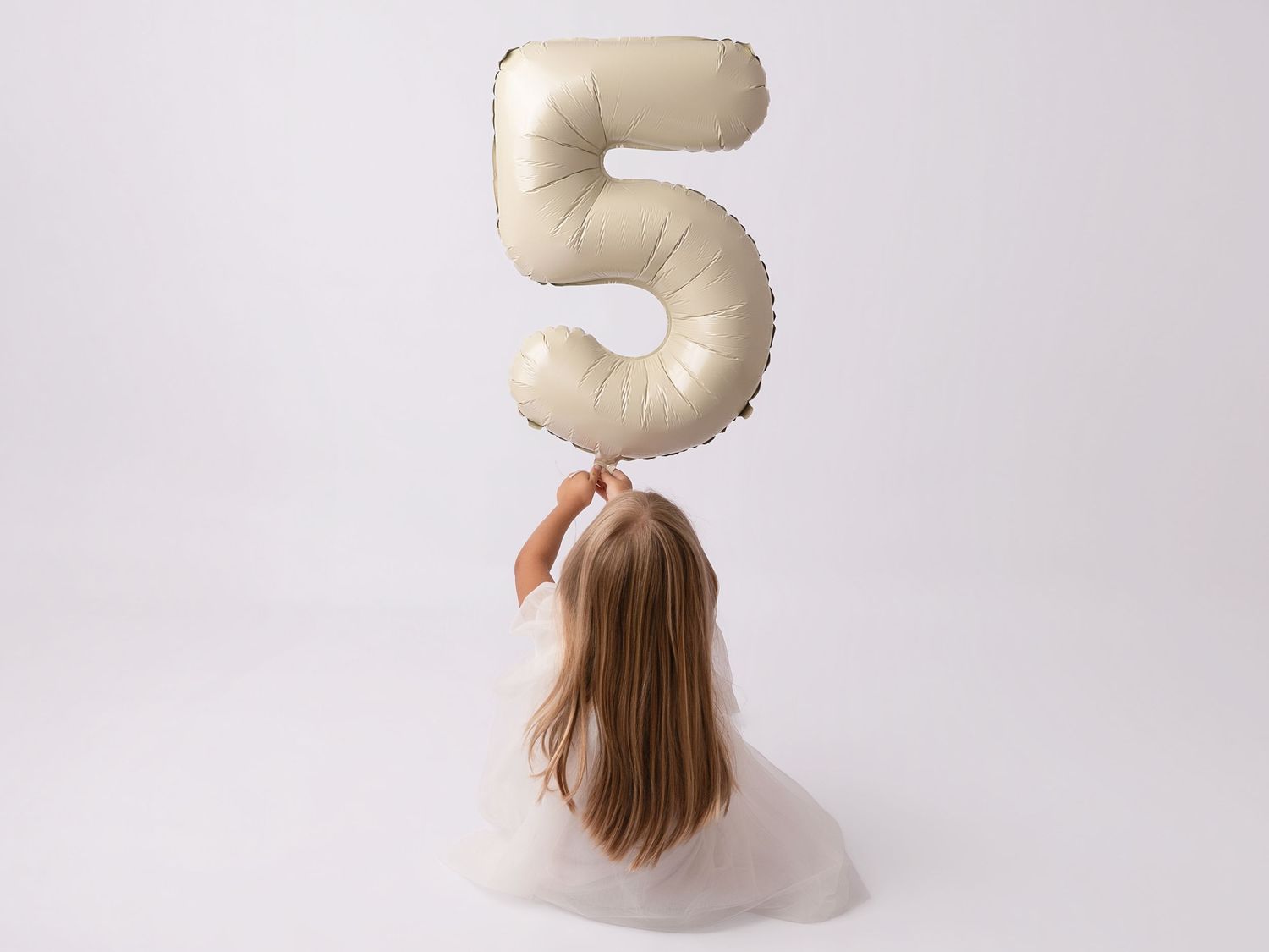 A young girl with long hair holds a large number 5 balloon, seen from behind, wearing a white tutu dress.