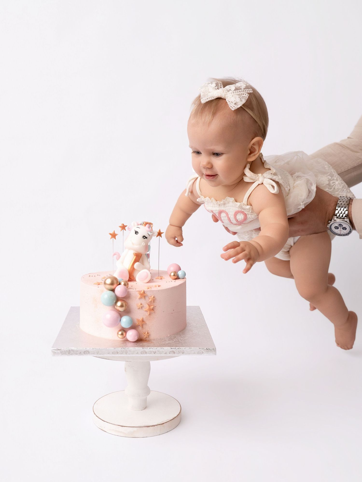 Baby girl in white romper reaching for a pink first birthday smash cake with unicorn topper on white background.