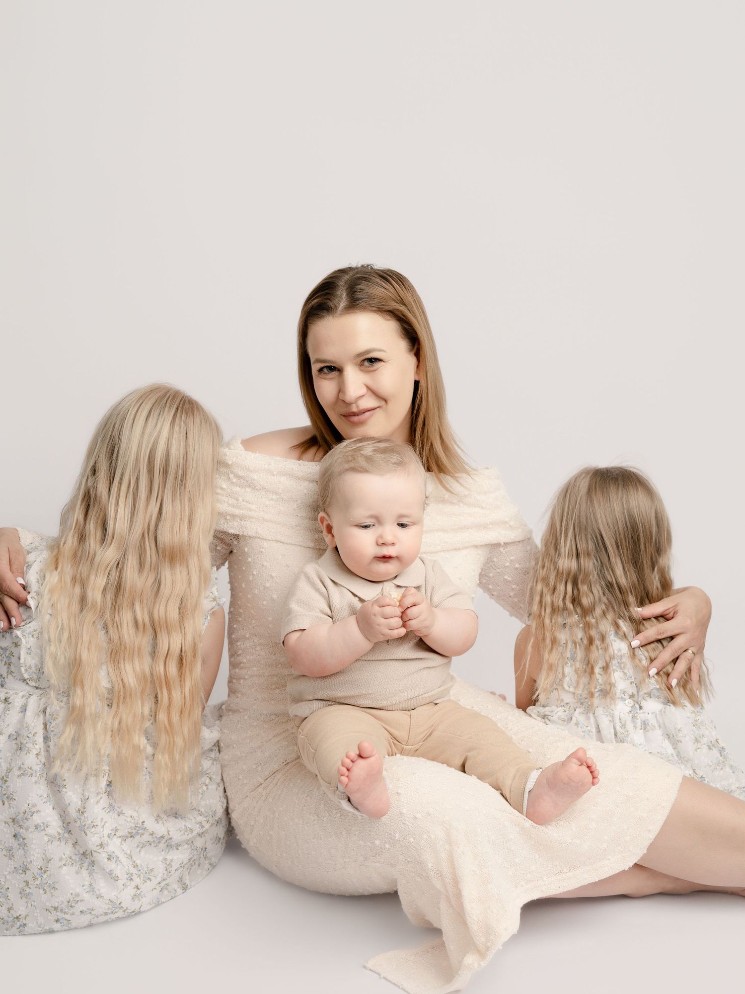 A mother in a white dress sits on the floor with her baby and two daughters facing away, in a soft studio setting.