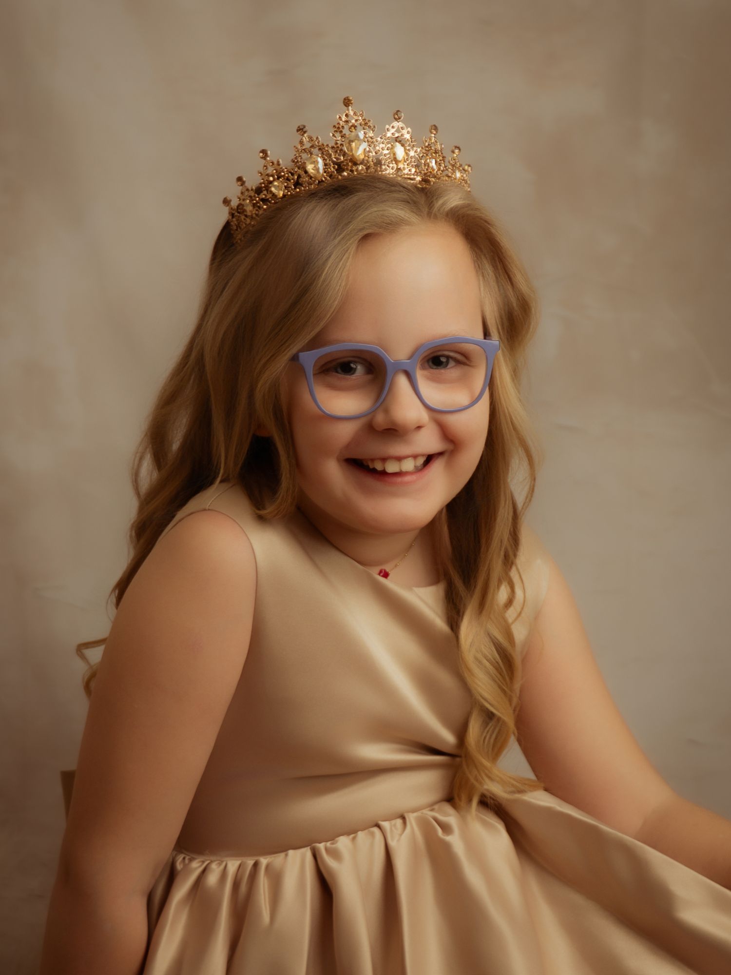 Smiling young girl wearing gold crown and blue glasses in elegant golden dress portrait.