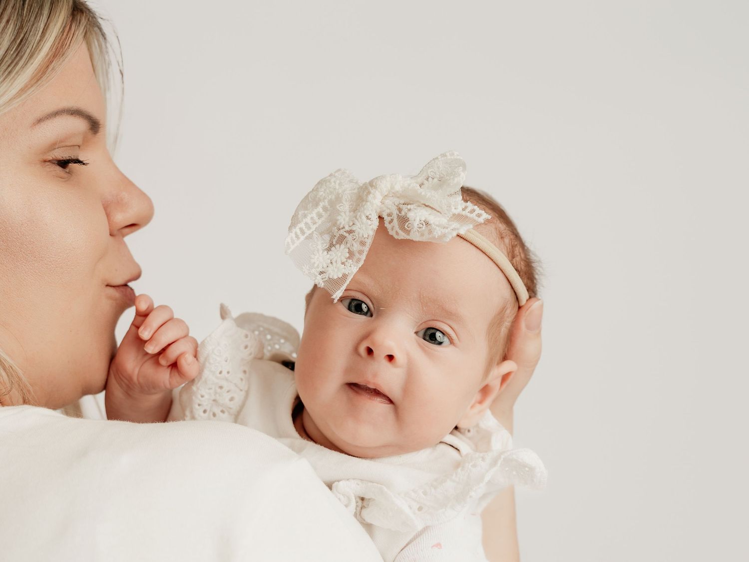 Blonde mother in white outfit holding alert newborn baby girl wearing lace bow headband.