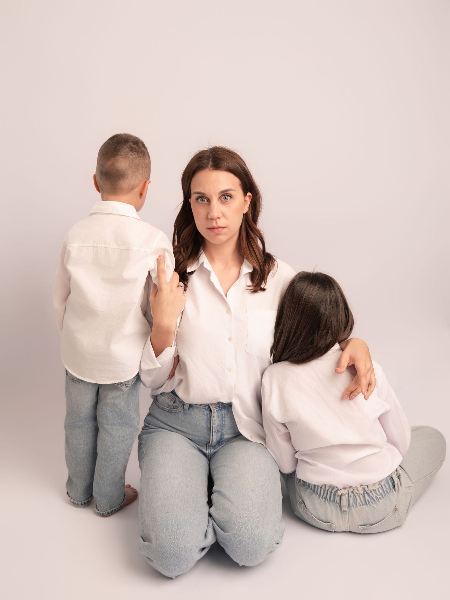 Mother kneeling with two children in matching white shirts and jeans in soft studio portrait.
