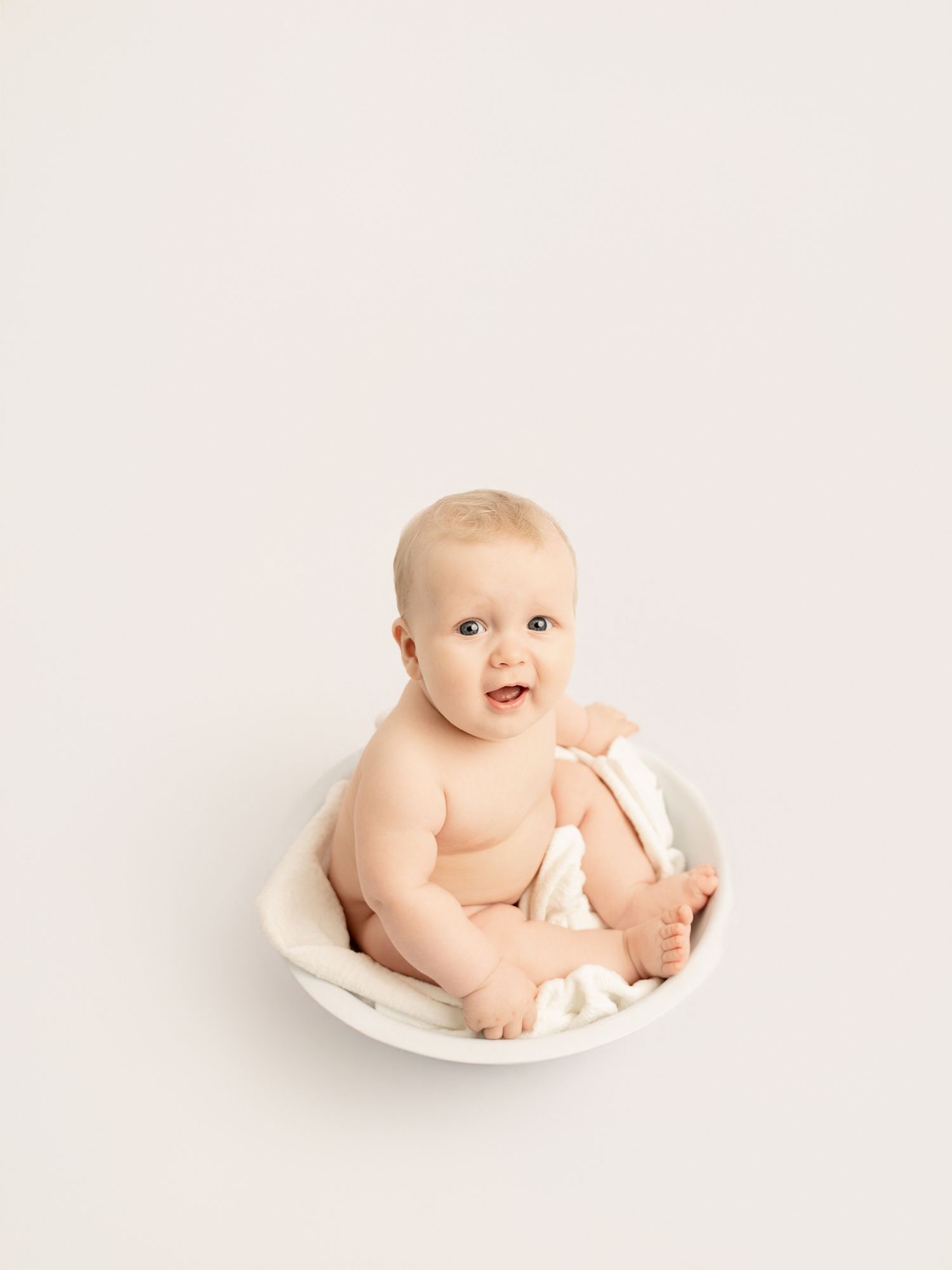 Sweet baby sitting in a round white bowl prop, smiling with blue eyes in a studio setting.