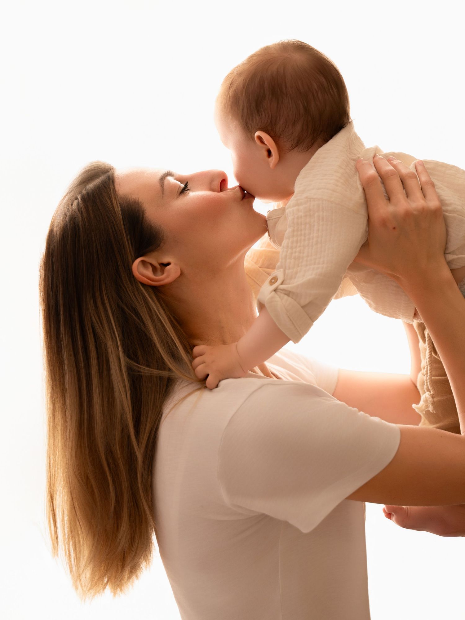 Mother lifting and kissing her baby in a bright studio, both wearing white outfits in a tender moment.