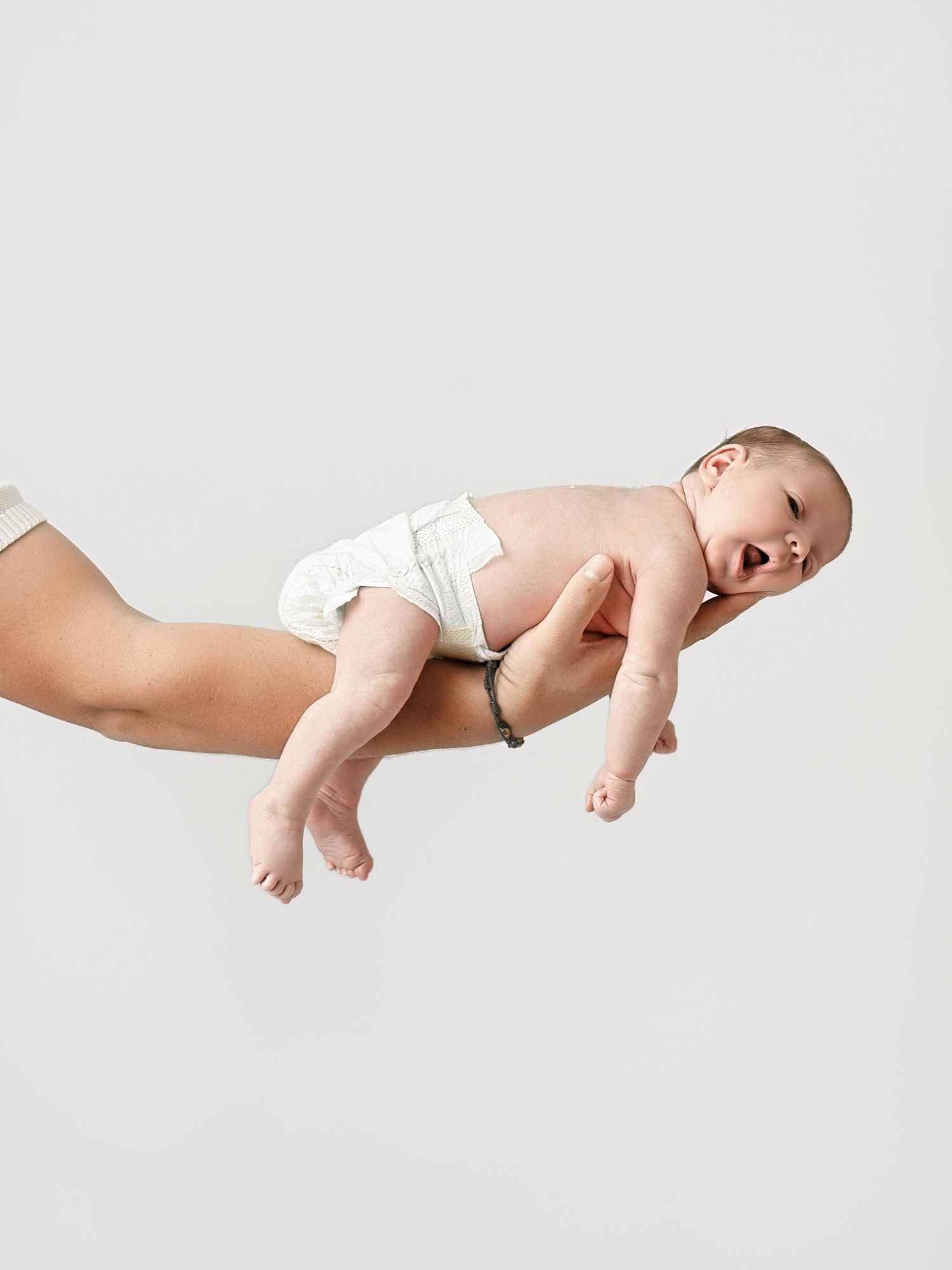 Newborn baby in white diaper being held horizontally by adult hands against a clean light gray background.