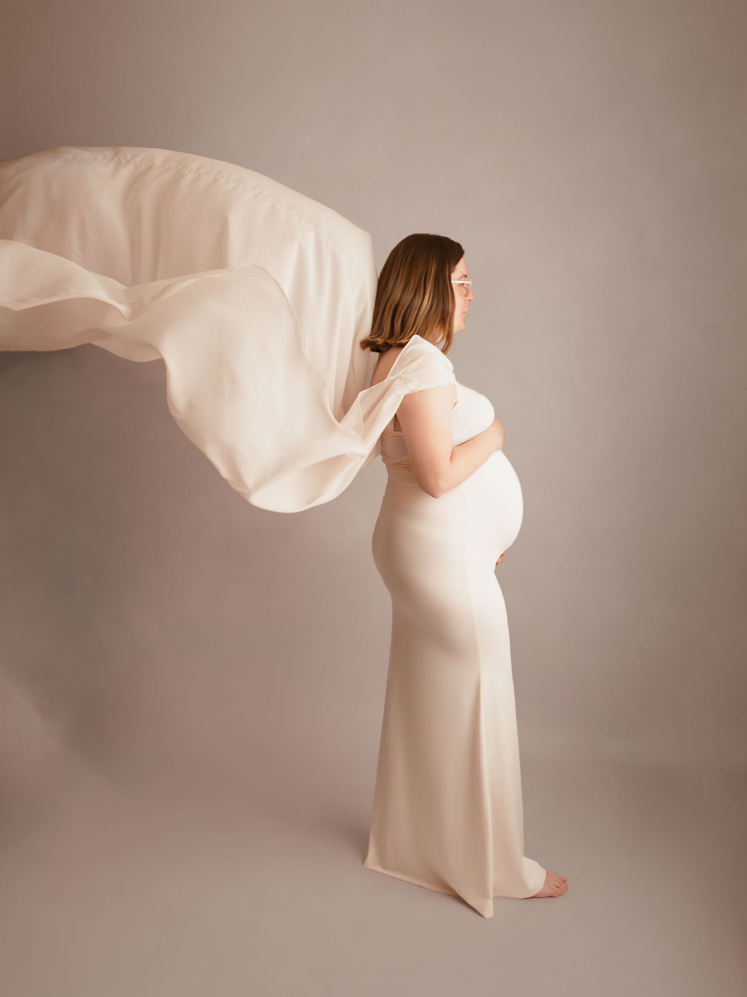 Pregnant woman in elegant cream gown with flowing fabric, posing sideways in a soft-lit studio maternity photoshoot.