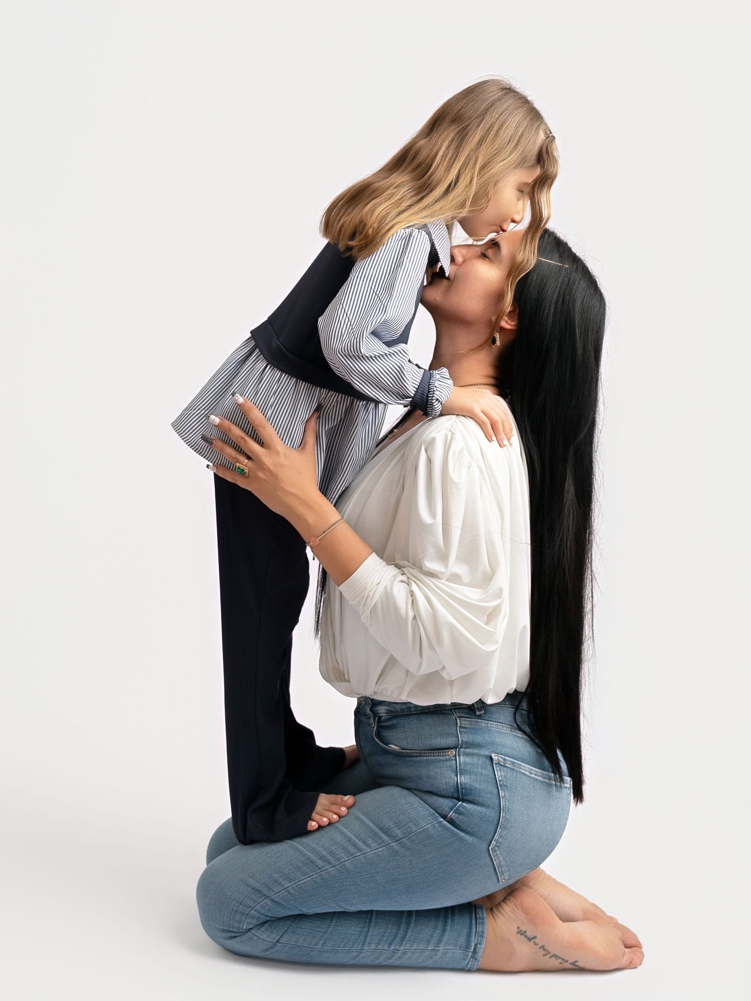 A mother kneeling on the floor lifts and kisses her young daughter in a bright studio photo.