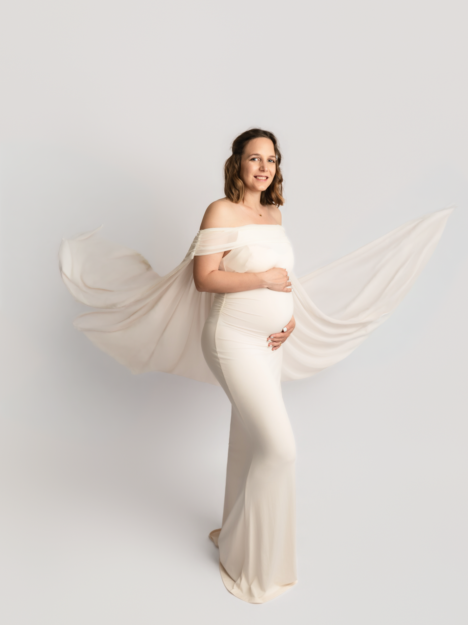 Smiling pregnant woman in cream off-shoulder gown with billowing chiffon cape in white studio.