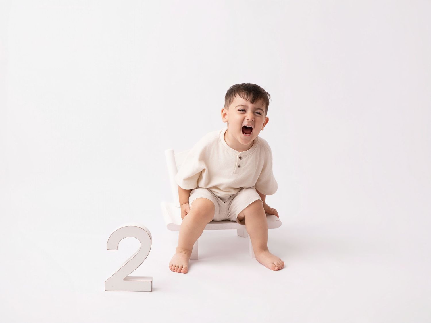 Toddler boy in beige romper sitting on small stool, laughing with number 2, white background.