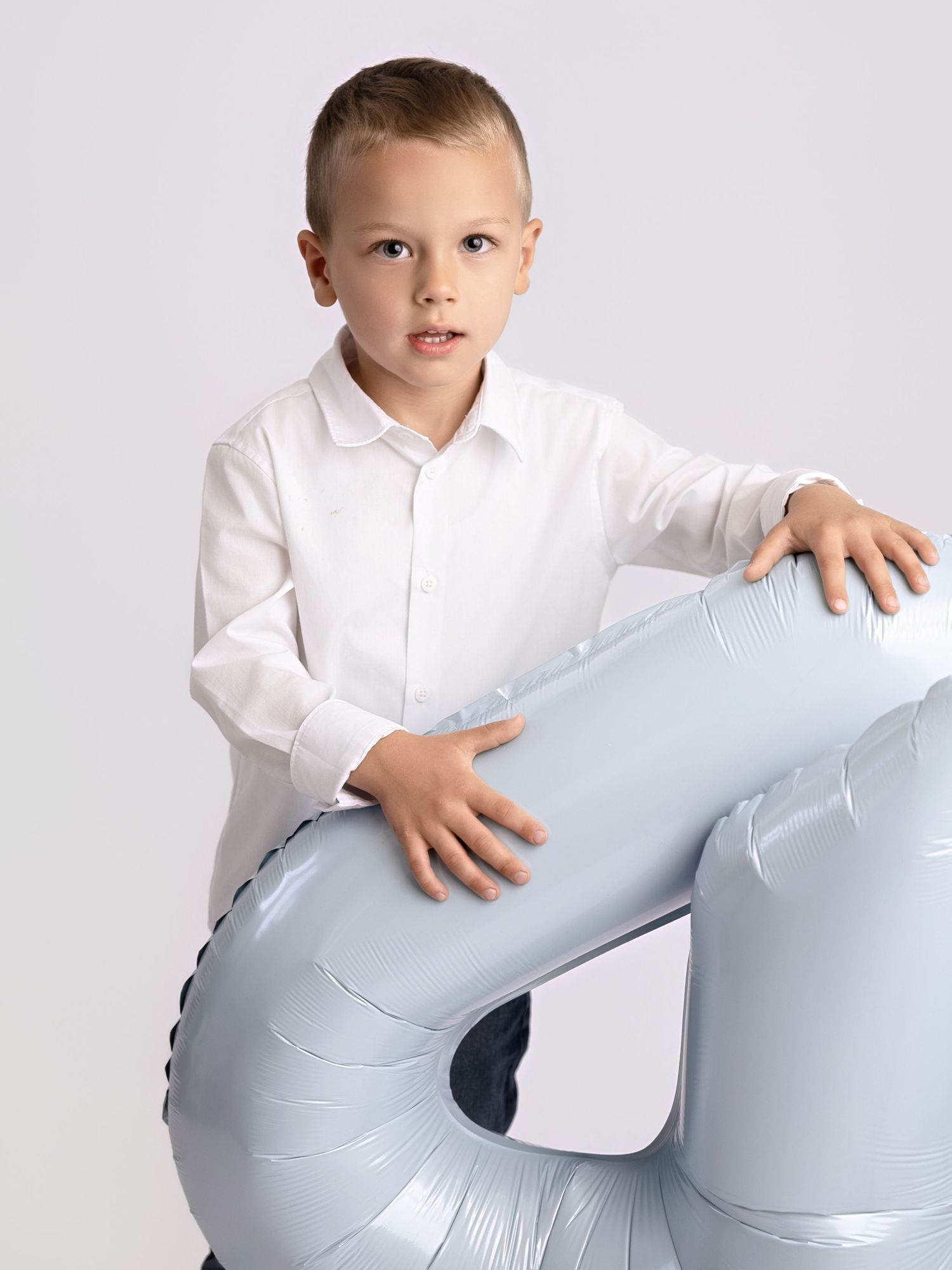 Young boy in white shirt hugs large silver number 4 balloon in white studio birthday portrait.