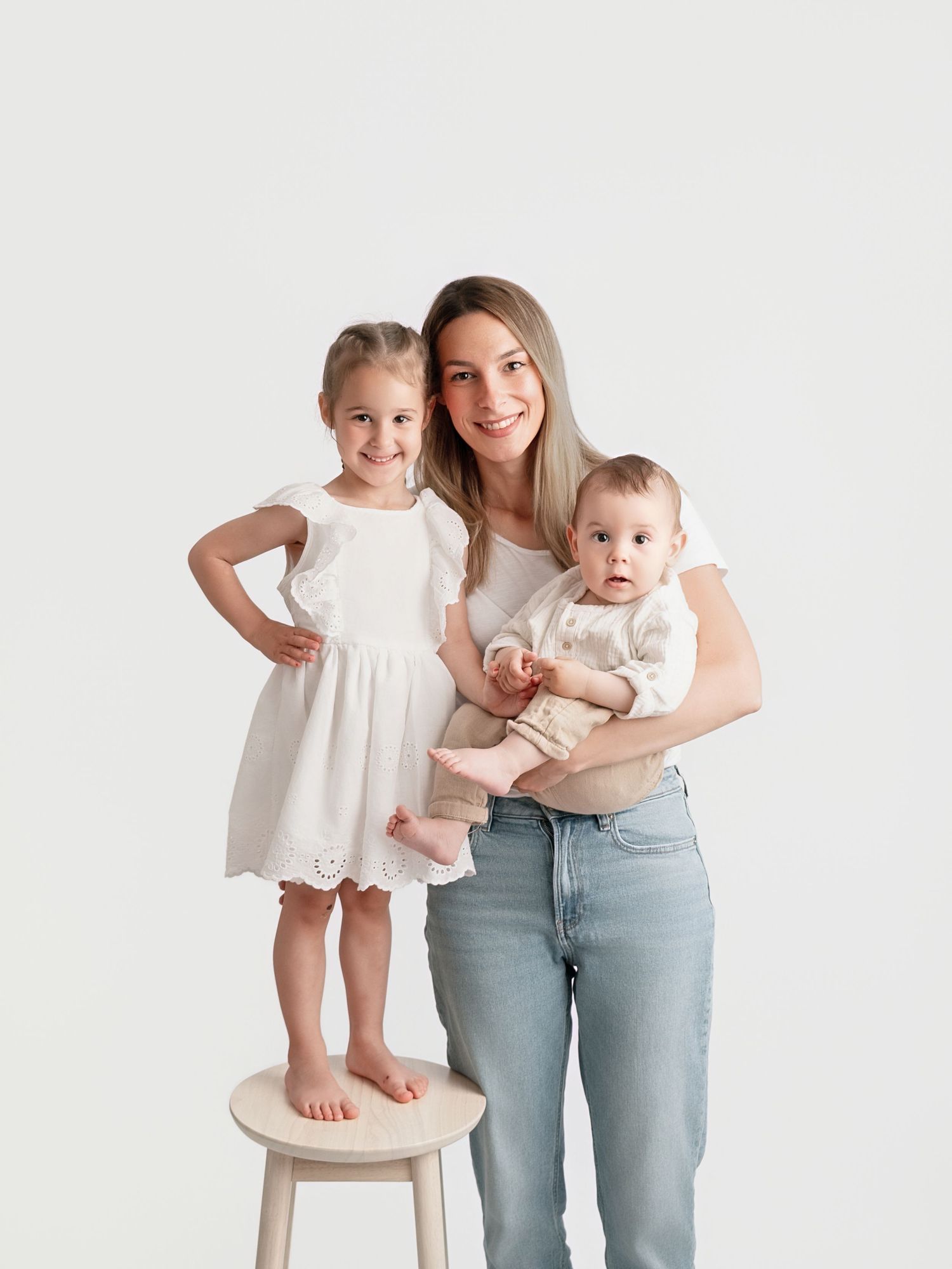 A woman in jeans holds a baby while a young girl in a white dress stands on a stool beside her.