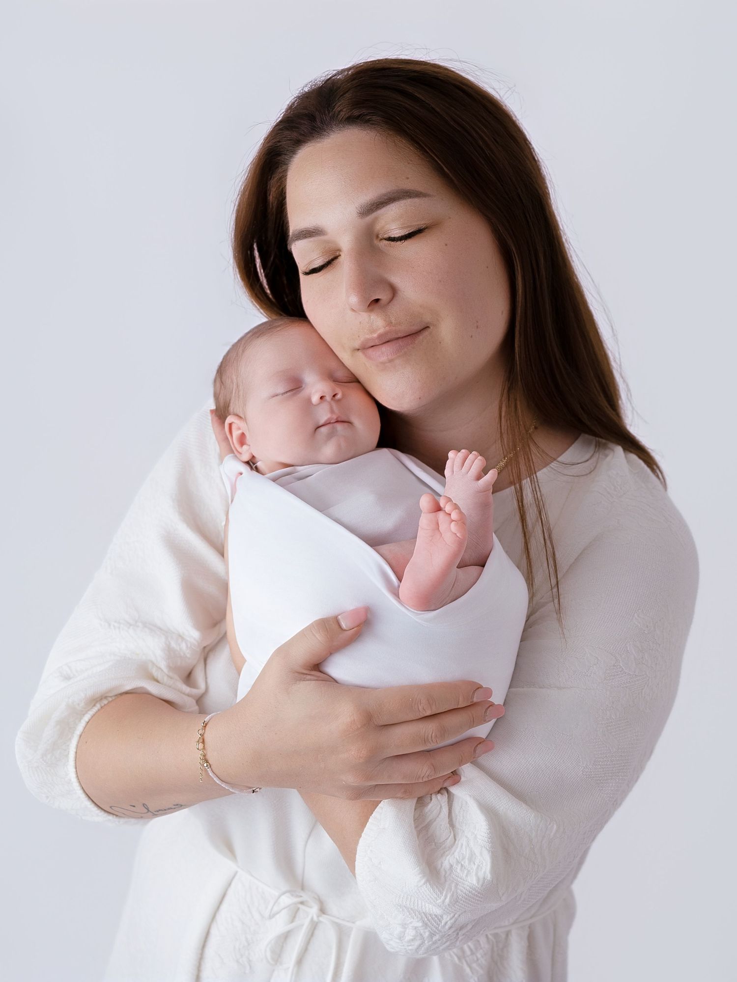 A mother in white gently cradles her sleeping newborn wrapped in a white swaddle against a soft gray background.