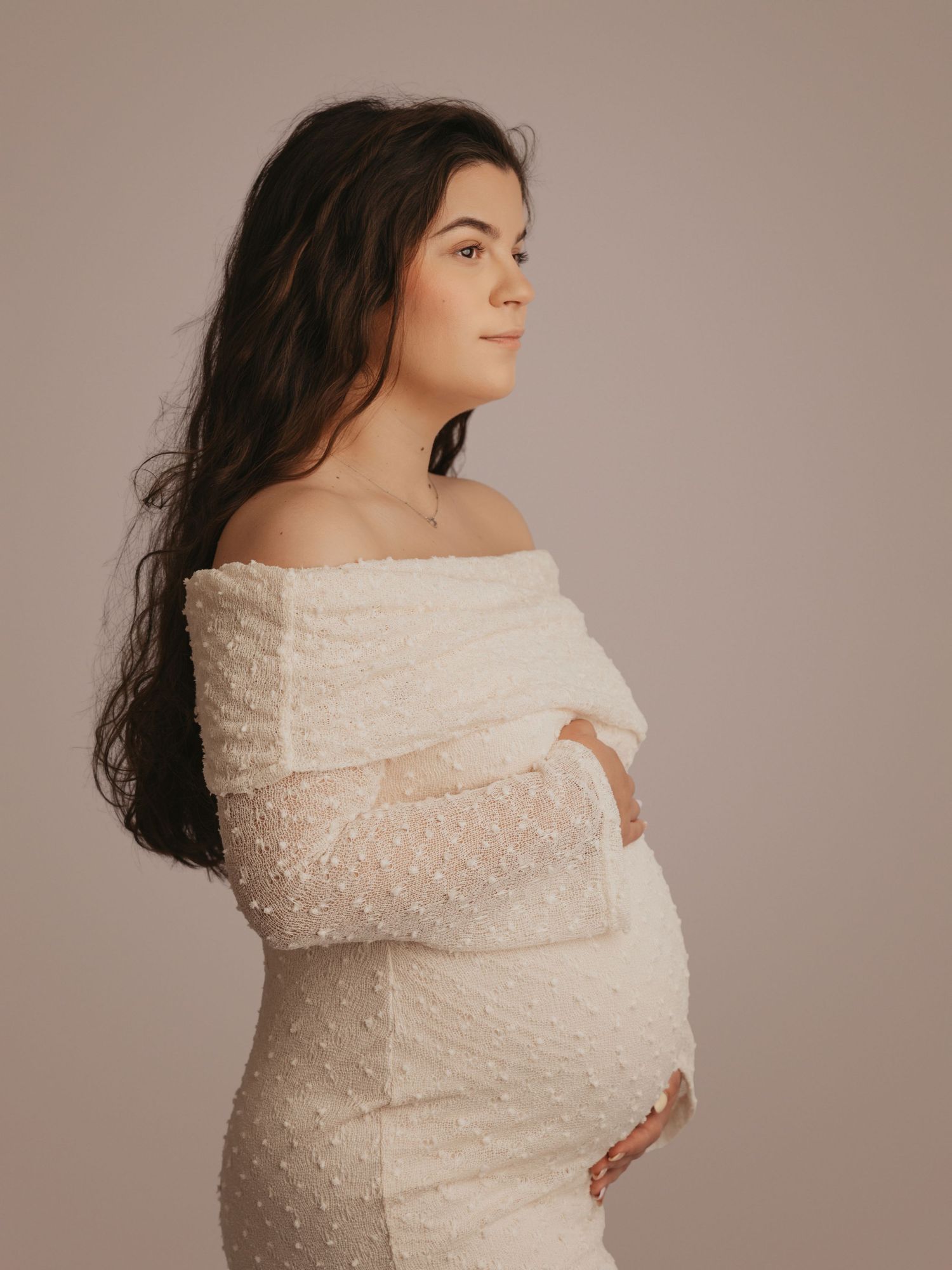 Pregnant woman in white off-shoulder beaded maternity dress with arms crossed over baby bump in studio.
