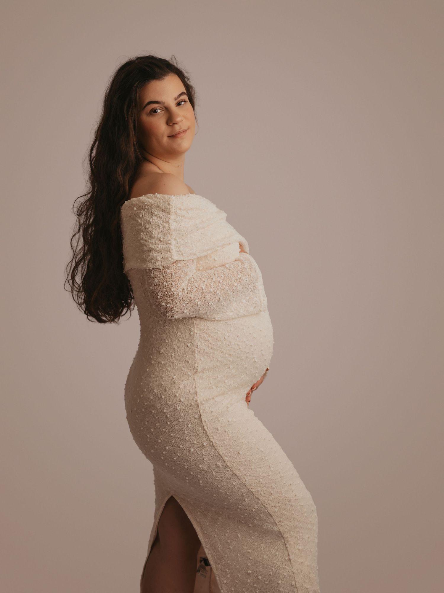 Pregnant woman in a fitted white pearl-embellished off-shoulder maternity dress posing in a bright studio.