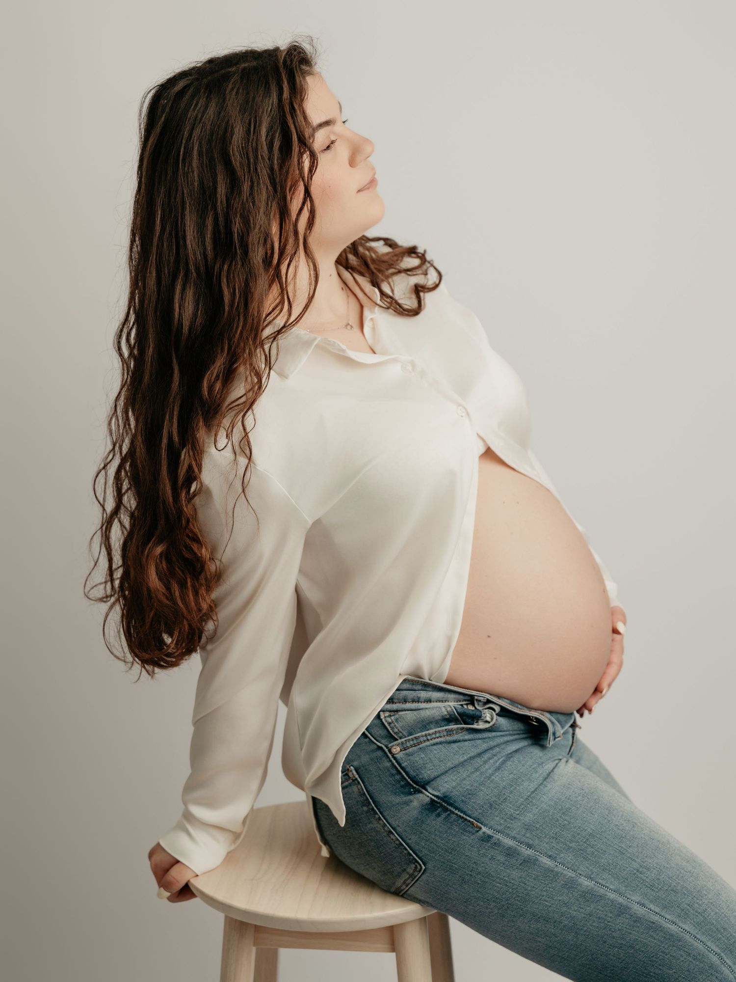 Pregnant woman sitting on stool in white shirt and jeans, cradling her bare baby bump in a studio setting.