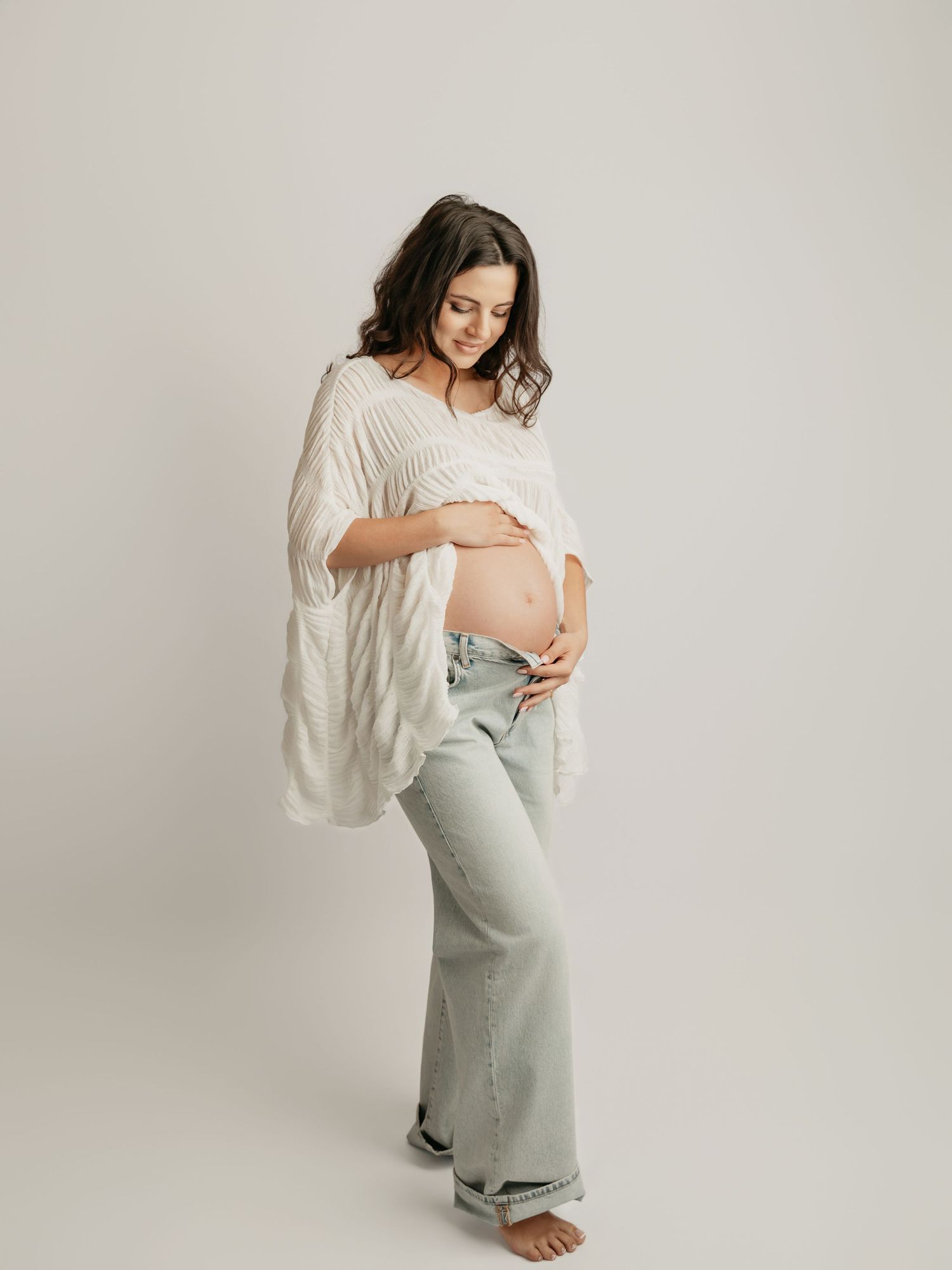 Full-length studio portrait of pregnant woman in white top and light jeans cradling bare bump.