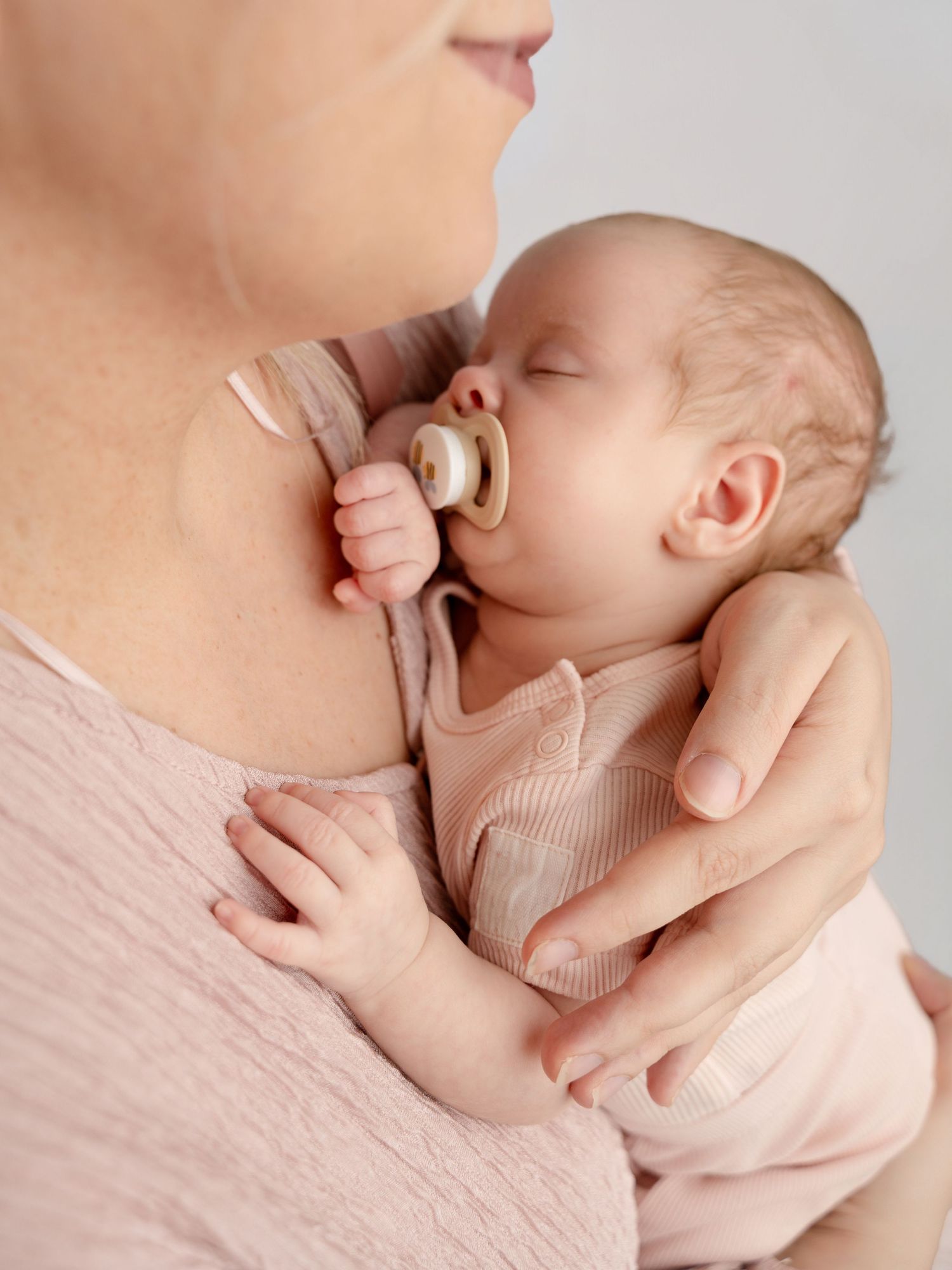 Close-up of newborn baby with pacifier sleeping against mother's chest in soft pink clothing.