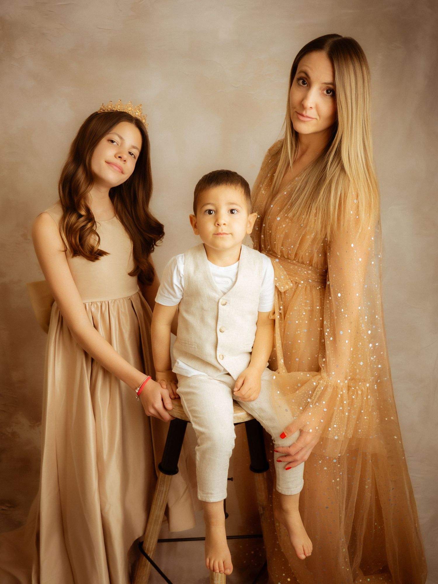 A mother in a gold gown poses with her two children in beige outfits for a studio portrait.