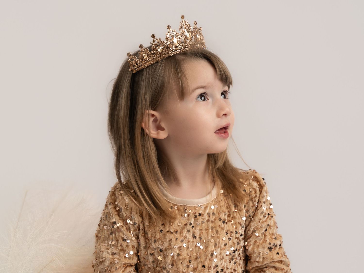 A young girl wearing a gold sequin dress and crown tiara looks upward in a soft, elegant portrait photo.