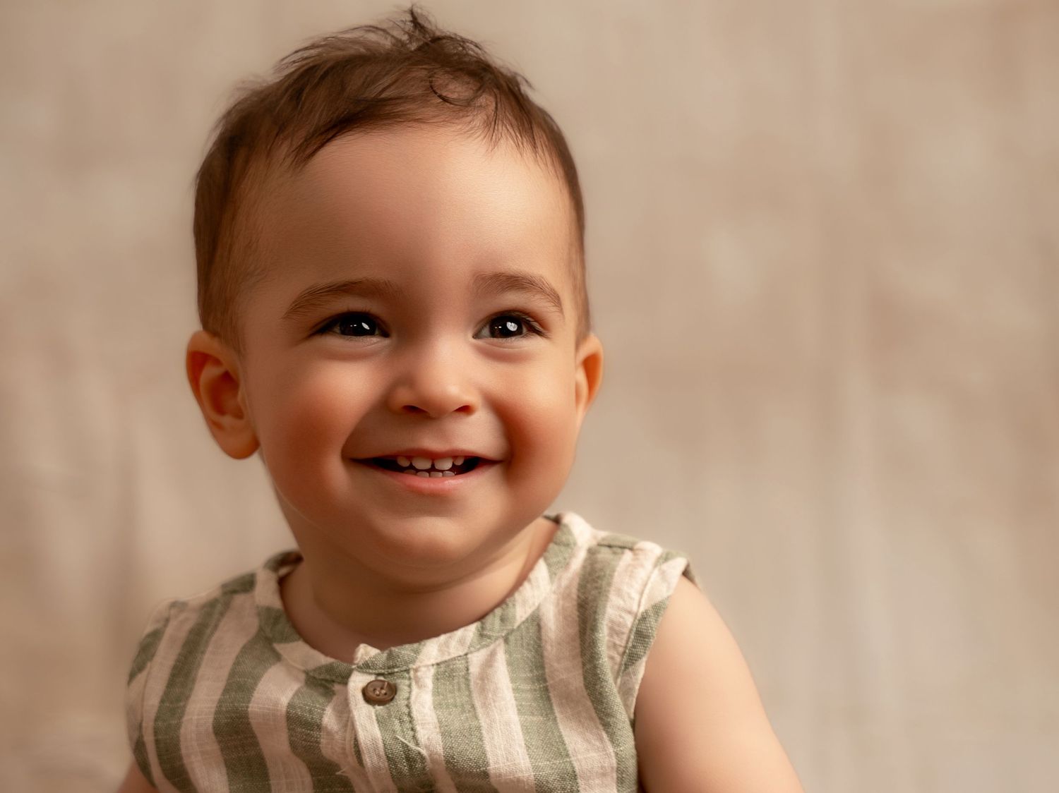 Smiling baby boy in striped green romper with brown hair, joyful expression, soft neutral background.