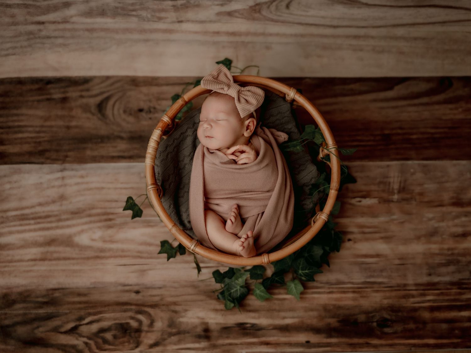 Newborn baby swaddled in beige wrap with bow headband sleeping in wicker basket surrounded by ivy on wood backdrop.