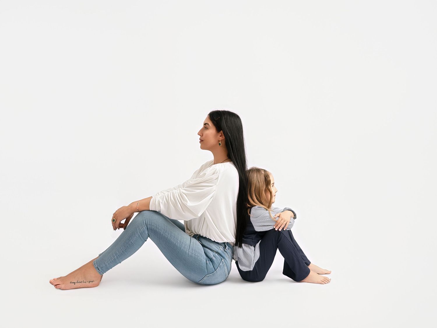 A mother and young daughter sit back-to-back on a white background, both wearing jeans and casual tops.